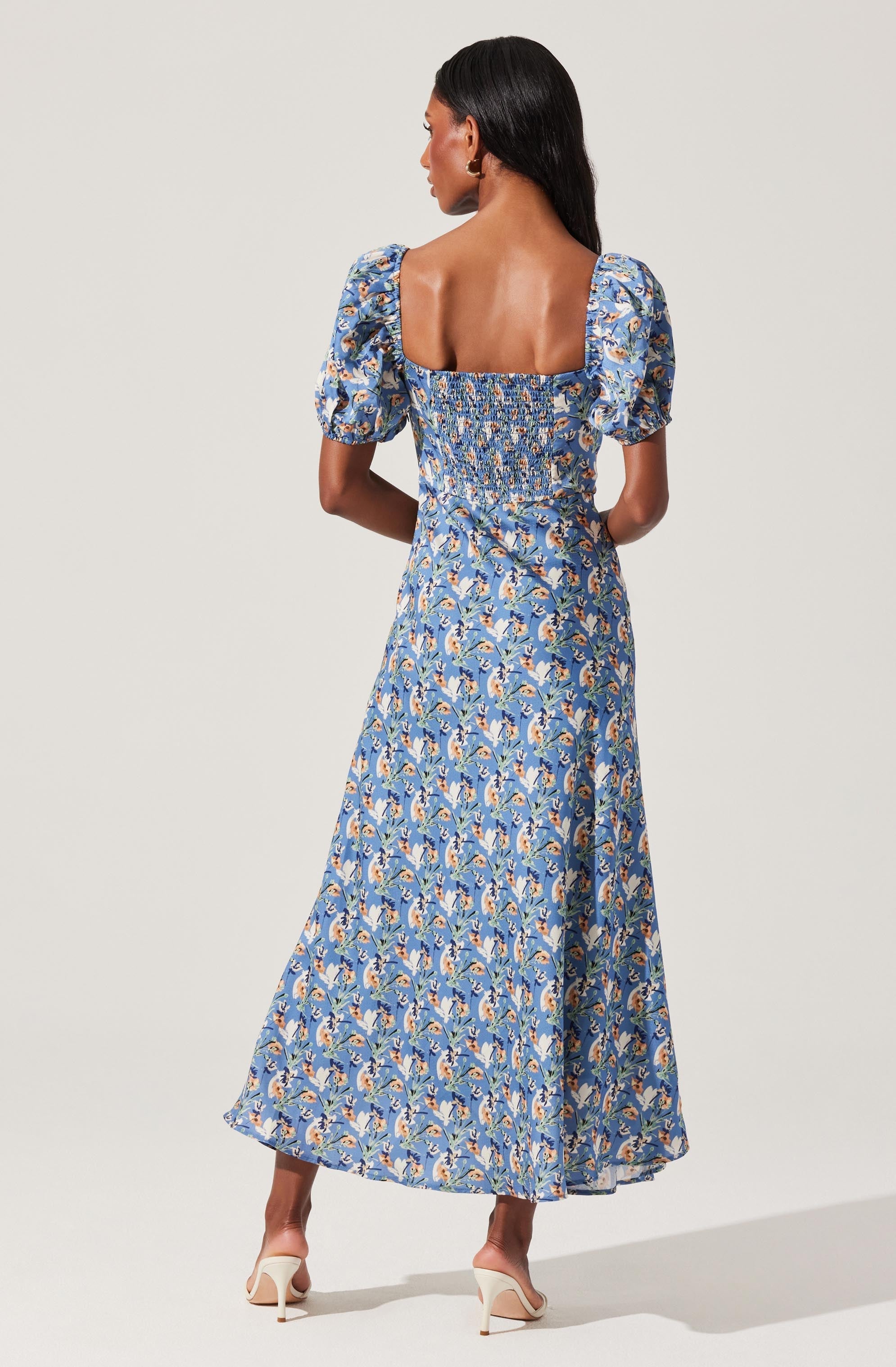 A woman is wearing a long, flowy blue dress with a floral pattern featuring shades of yellow, white, and pink. The dress has puffed short sleeves with elasticized cuffs, and a square neckline with a smocked bodice. She is also wearing beige high-heeled shoes.