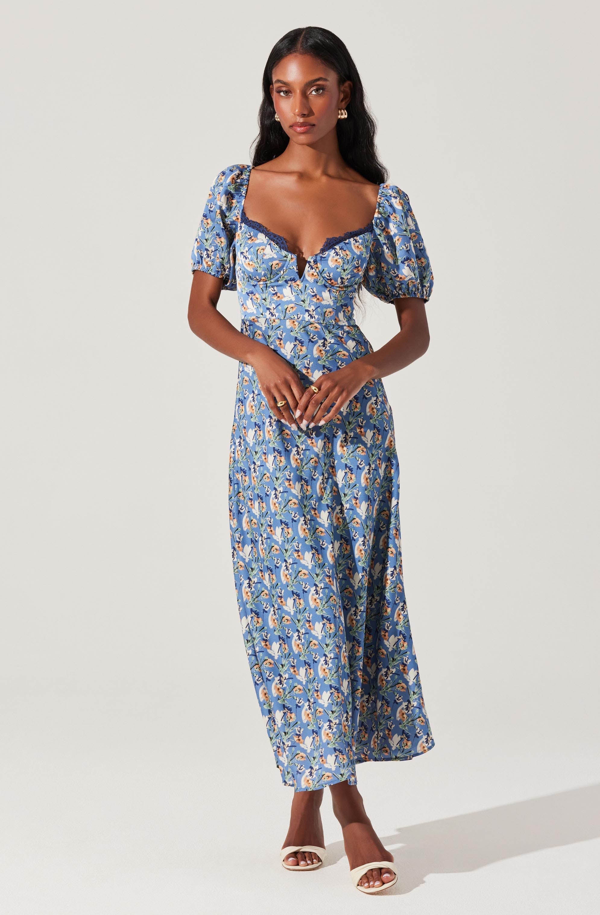 A woman is wearing a long, blue floral dress with a fitted bodice and a sweetheart neckline accented with dark blue lace trim. The dress has short, puffy sleeves and a flowing, ankle-length skirt. She is also wearing open-toed, white high-heeled sandals.