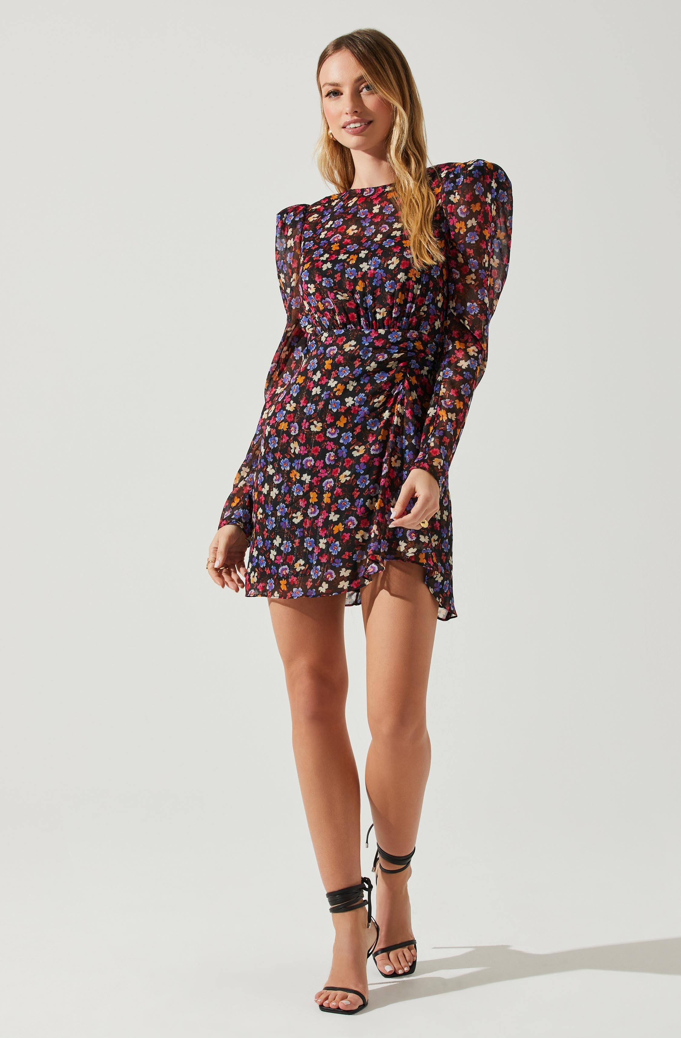 A woman is wearing a black dress with a colorful floral pattern, featuring mostly pink, purple, and blue flowers. The dress has long, sheer sleeves with puffed shoulders and a high neckline. The skirt of the dress is slightly ruffled and falls above the knees. She is also wearing black, strappy high-heeled sandals that wrap around her ankles.