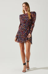 A woman is wearing a black dress with a colorful floral pattern, featuring mostly pink, purple, and blue flowers. The dress has long, sheer sleeves with puffed shoulders and a high neckline. The skirt of the dress is slightly ruffled and falls above the knees. She is also wearing black, strappy high-heeled sandals that wrap around her ankles.