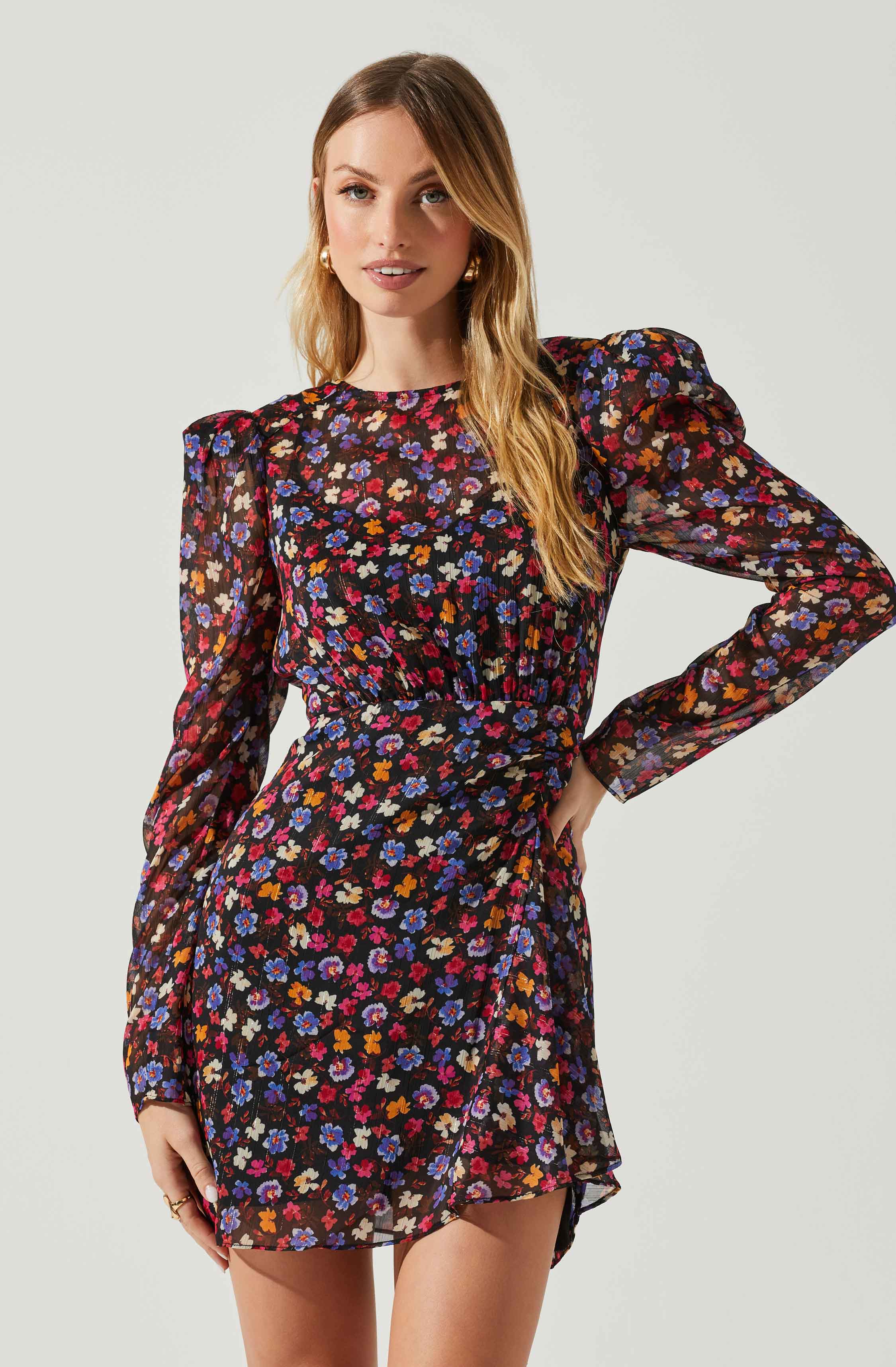 A woman is wearing a long-sleeved, black dress with a colorful floral pattern featuring red, purple, orange, and cream flowers. The dress has a fitted waist and a slightly gathered skirt.