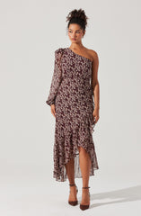 A woman is wearing a long, flowy dress with a high-low hemline. The dress features a dark background with a small, light-colored floral print. It has an asymmetrical, off-the-shoulder neckline, with one long puffed sleeve on the left side and sleeveless on the right. The dress is fitted at the waist and cascades down in ruffled layers, ending above the ankles in the front and extending to the ankles at the back. She is wearing brown high-heeled shoes.