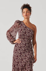 A woman is wearing a dress with a dark background and a light floral pattern. The dress has an asymmetrical neckline, exposing one shoulder, and features a single puff sleeve on the left side. The fabric appears lightweight and sheer, especially on the sleeve. The dress is fitted at the waist and has a ruffled, asymmetrical hemline that falls to mid-length.