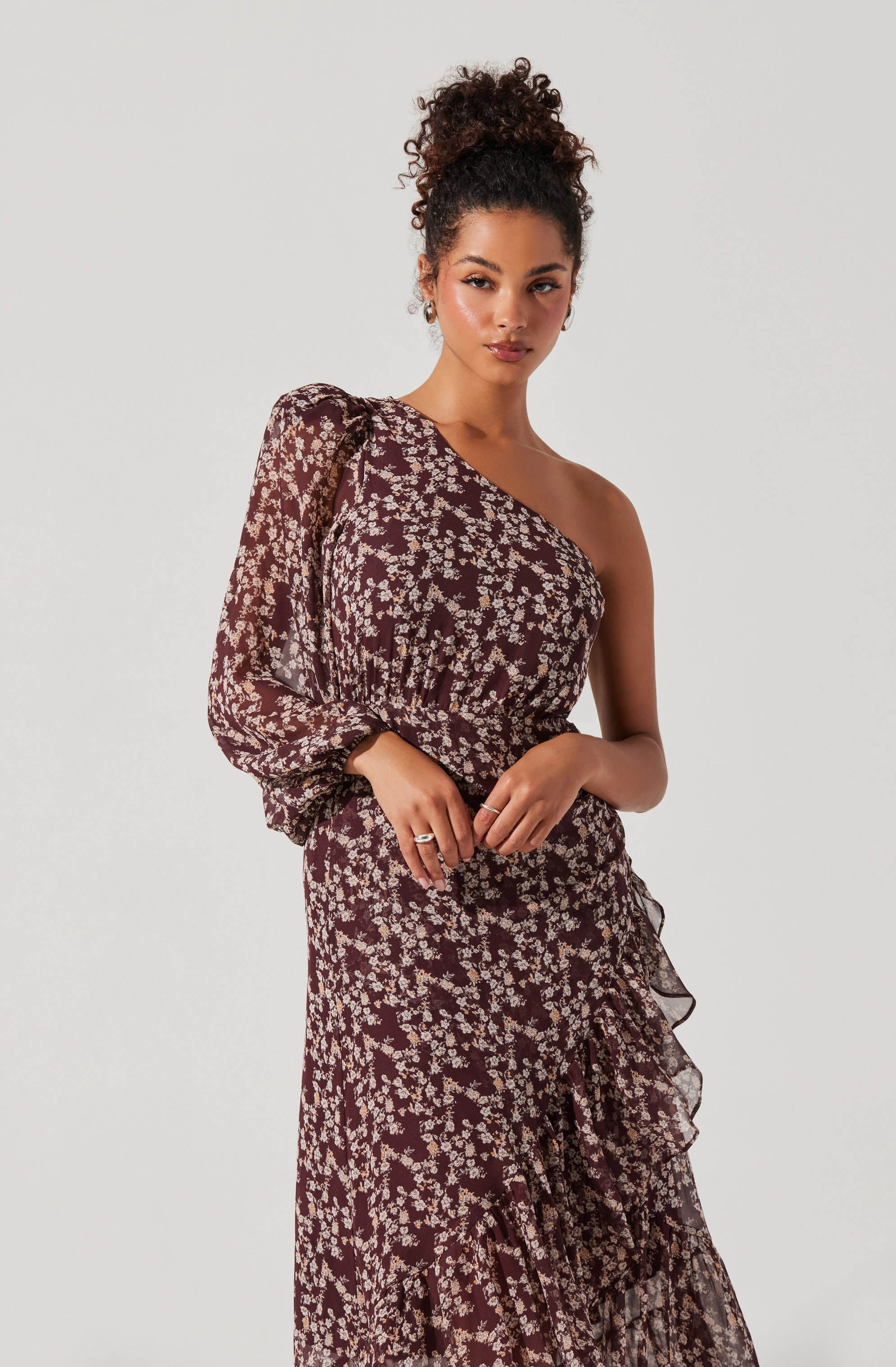 A woman is wearing a dark-colored dress with a floral pattern in light beige or cream. The dress features a one-shoulder design, with a long, sheer sleeve on the left side that is slightly puffed at the shoulder. The fabric of the dress appears lightweight with a ruffled detail running diagonally across the skirt. The overall look is elegant and stylish.