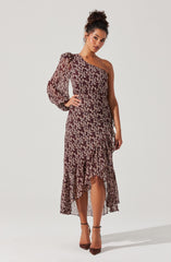 A woman is wearing a dress with a one-shoulder design. The dress has a fitted bodice and a high-low hemline with ruffled detailing. It features a floral pattern in shades of dark red, pink, and cream on a dark background. The dress has long, sheer, puffed sleeves on one side, while the other shoulder remains bare.