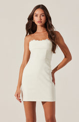 A woman is wearing a strapless, fitted white dress that has a scalloped lace trim along the top edge. The dress features a structured bodice with vertical seam detailing, and the length is above the knees. She accessorizes with gold hoop earrings and has long, wavy brown hair.