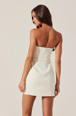 A woman with long, straight brown hair is wearing a strapless, cream-colored mini dress with a fitted bodice and a zipper closure at the back. The dress has a smooth, slightly shiny fabric and a simple, elegant design.