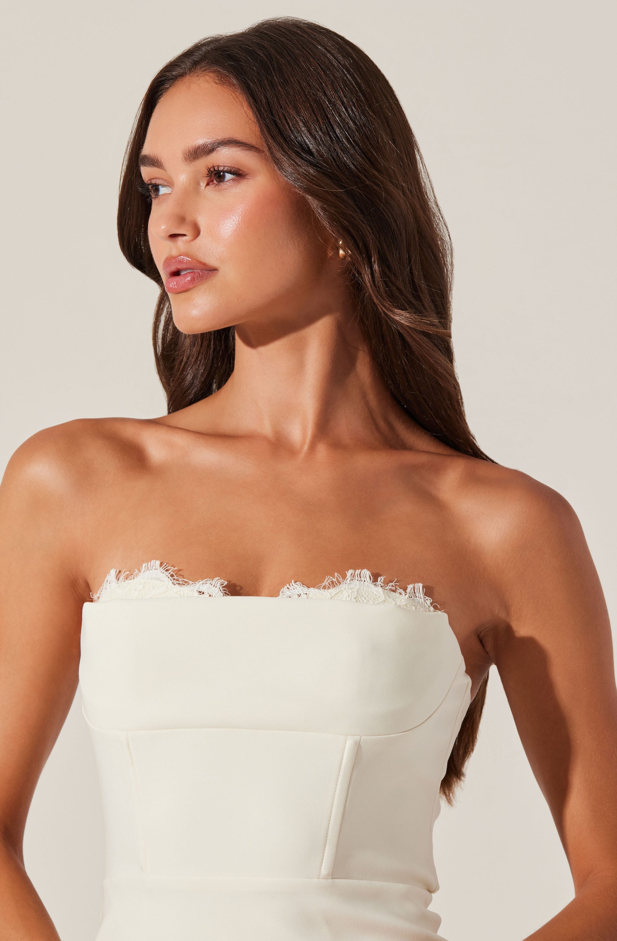 A woman is wearing a strapless white dress with a straight neckline accented by delicate lace trim. The dress appears fitted at the bodice with seam detailing, and she has a natural, elegant look with long, dark brown hair styled in loose waves.