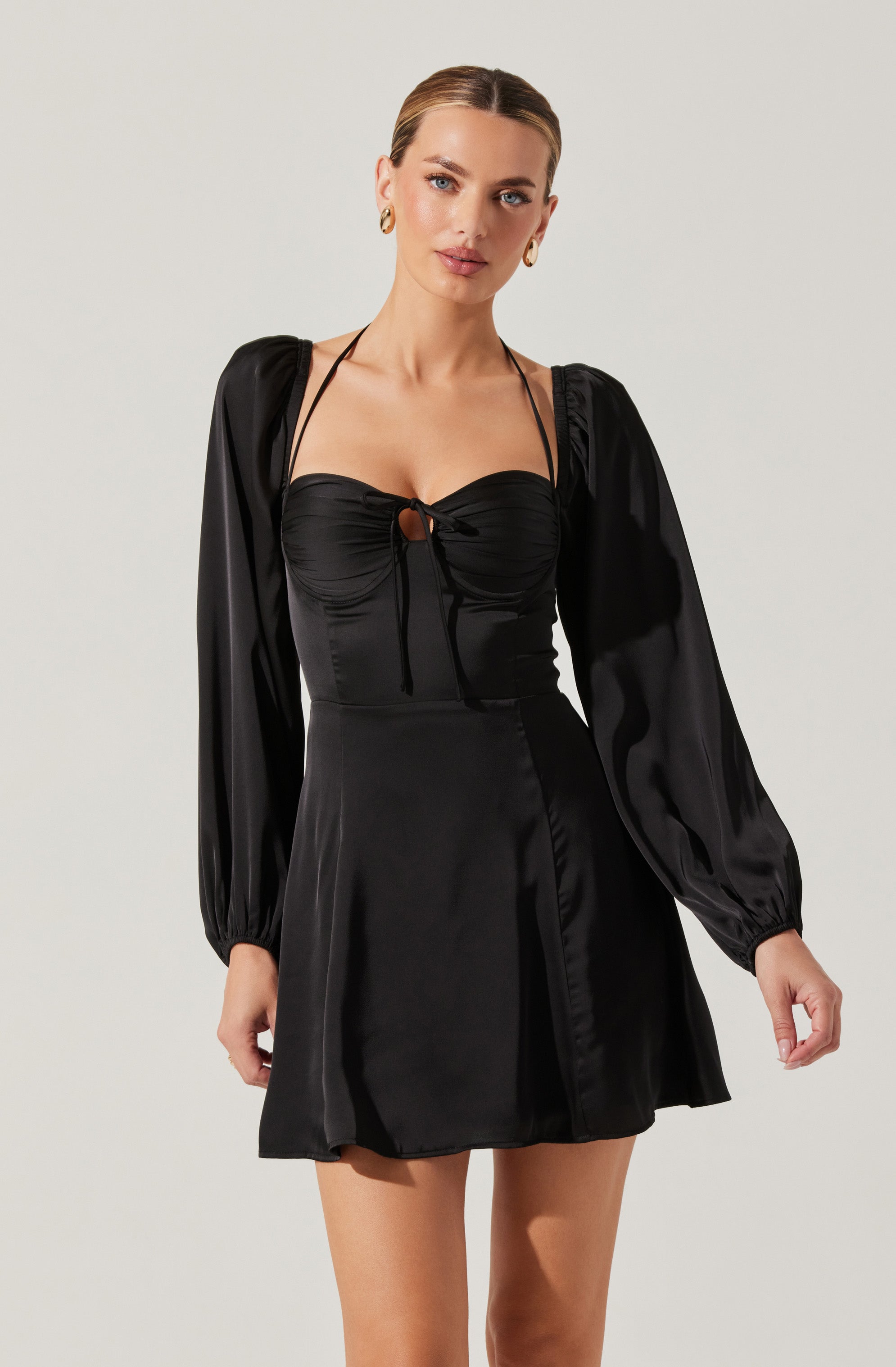 A woman is wearing a black satin dress featuring long, puffed sleeves with elastic cuffs, a fitted bodice with ruched detailing at the bust, and a small keyhole opening with a tie closure at the center of the chest. The dress has a slightly flared skirt that falls above the knees. She accessorizes with gold hoop earrings.