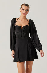 A woman is wearing a black satin dress featuring long, puffed sleeves with elastic cuffs, a fitted bodice with ruched detailing at the bust, and a small keyhole opening with a tie closure at the center of the chest. The dress has a slightly flared skirt that falls above the knees. She accessorizes with gold hoop earrings.