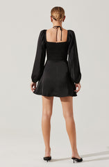 A woman is wearing a black dress with long, puffed sleeves made of shiny fabric. The dress has a square neckline and a fitted, smocked bodice, with a flared skirt that ends above the knees. The back of the dress features a tie at the neck and an open square cutout at the top of the back. She is also wearing black open-toe mules with a small heel.