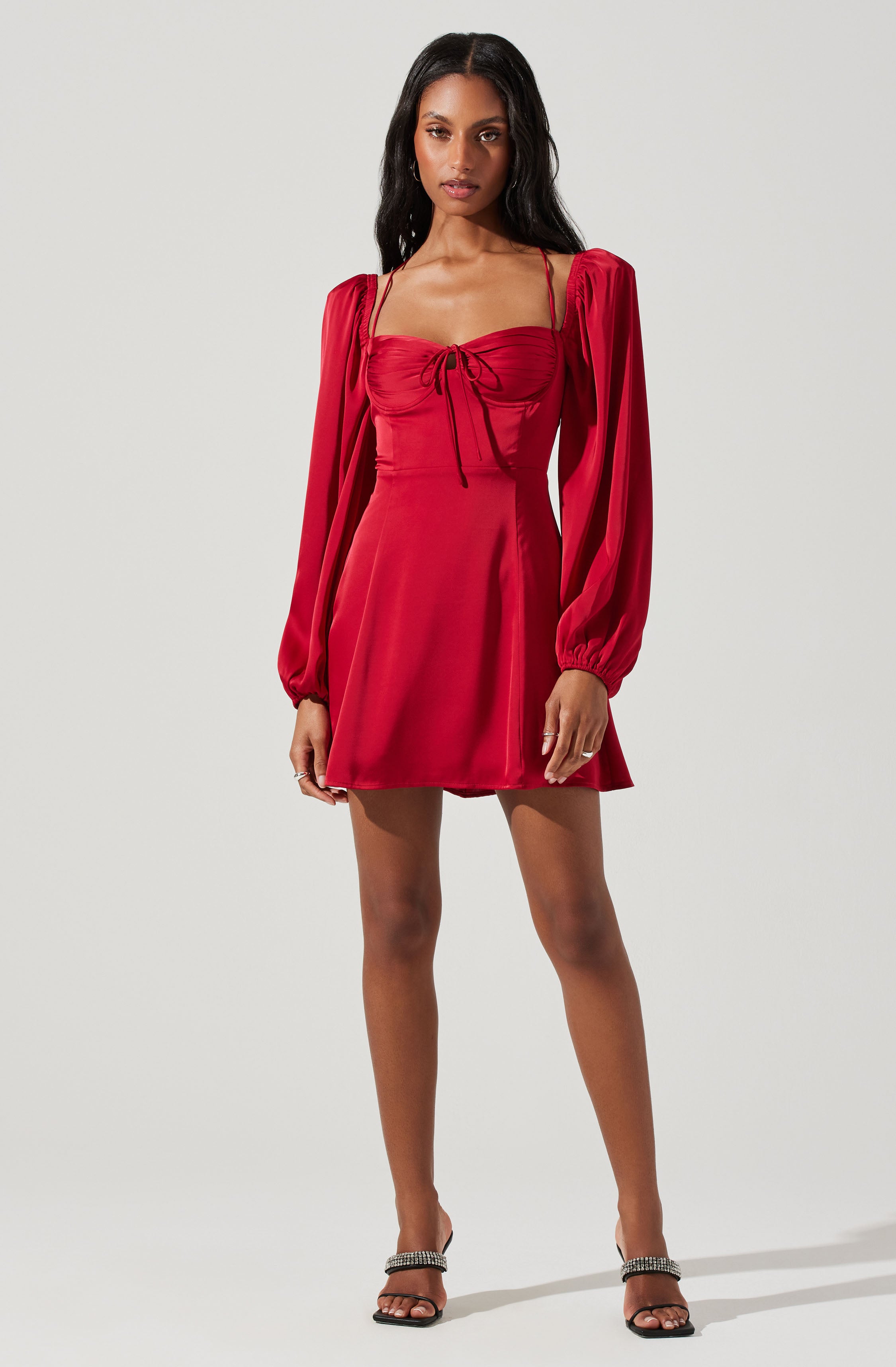 A woman is wearing a red dress with a fitted bust and a bow detail at the front. The dress has long, puffed sleeves with elastic cuffs and thin spaghetti straps. The skirt is short and flares out slightly. She pairs the dress with black high-heeled sandals that have embellishments on the straps.
