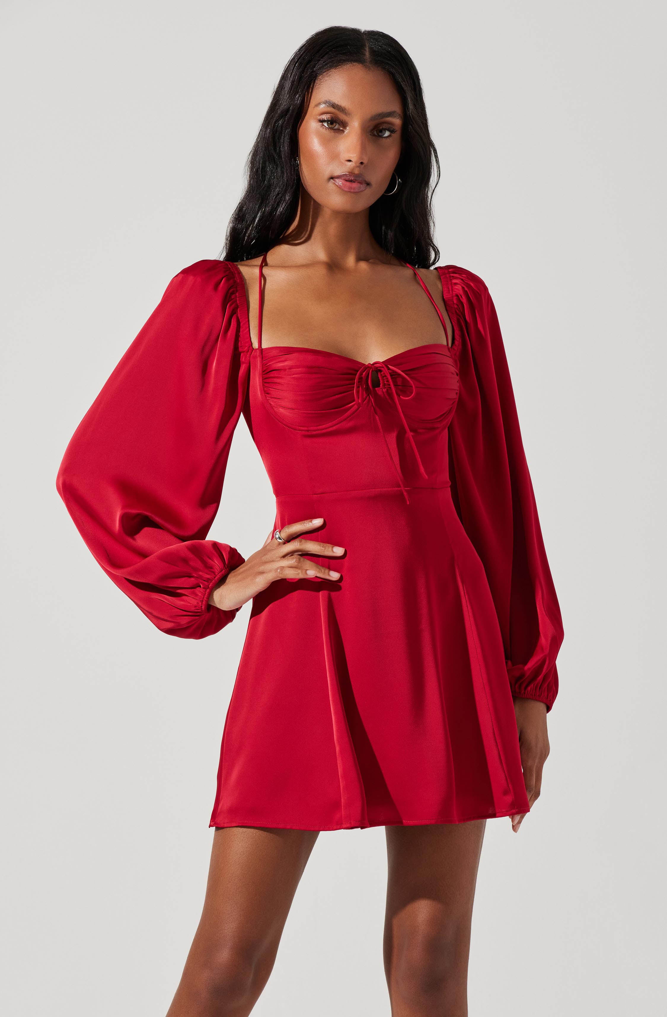 A woman is wearing a red dress with puffed, long sleeves that are gathered at the wrists, and shoulder straps. The dress features a ruched bodice with a gathered detail and a bow tie at the center, and the skirt is slightly flared with vertical seams running down the front.