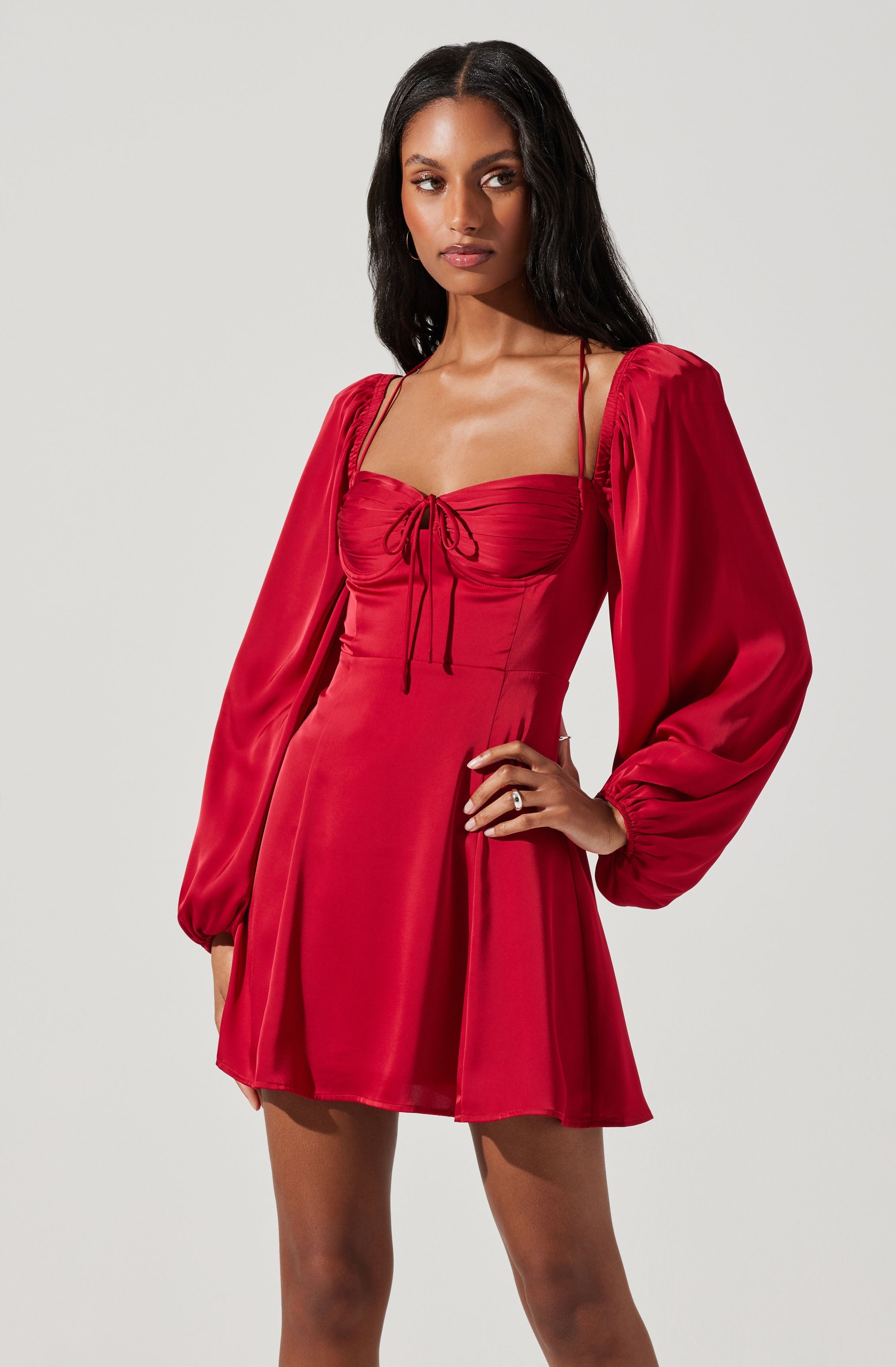 A woman is wearing a red dress with a fitted bodice and a short, flared skirt. The dress features long, puffed sleeves gathered at the wrists and thin shoulder straps. The bodice has a ruched detail at the front with a small bow tied in the center.