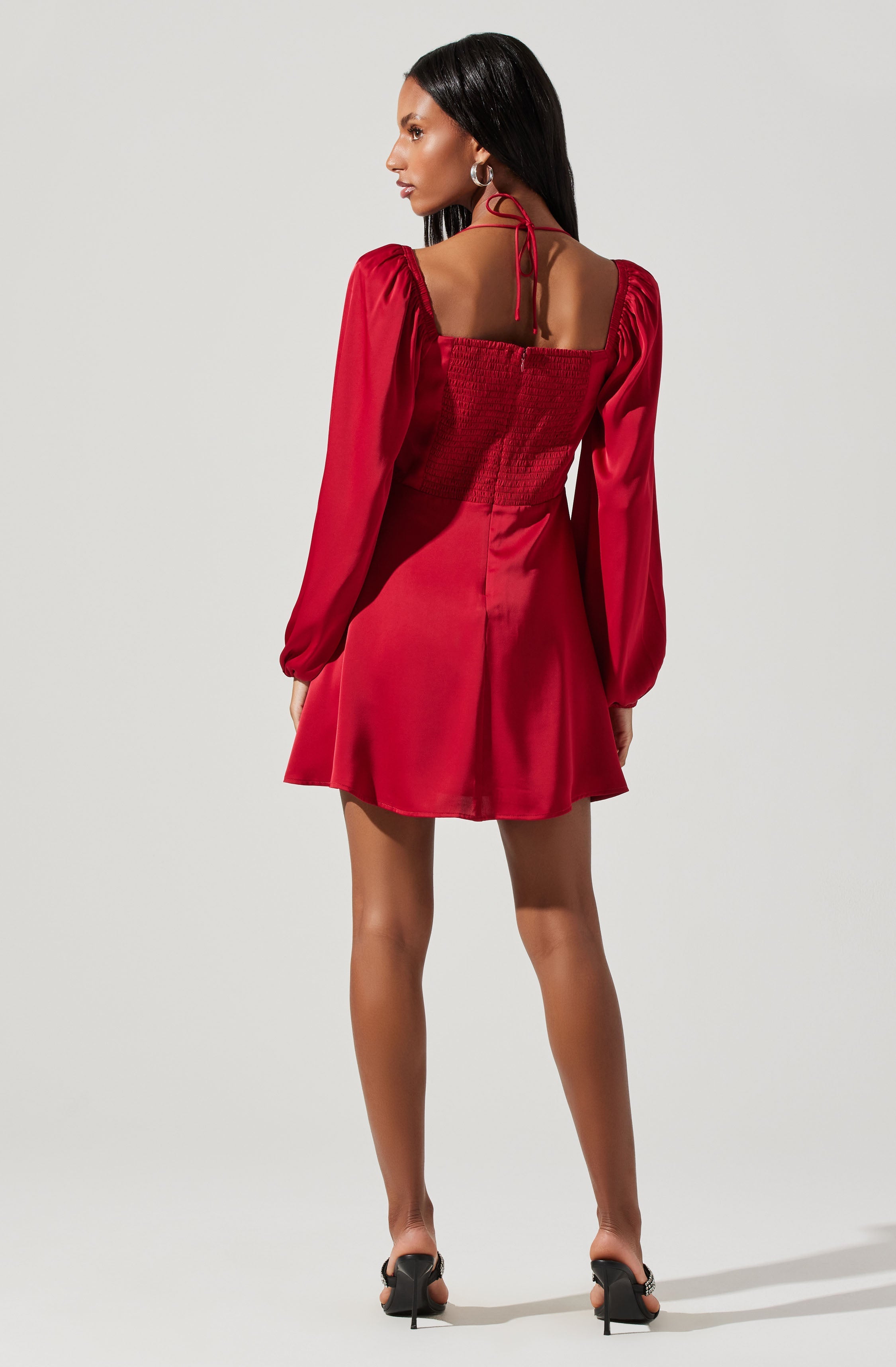 A woman is wearing a red dress with long, puffed sleeves and a square neckline with shoulder straps tied in a bow. The bodice features smocking and the skirt is short and flared. She is also wearing black high-heeled sandals and hoop earrings.