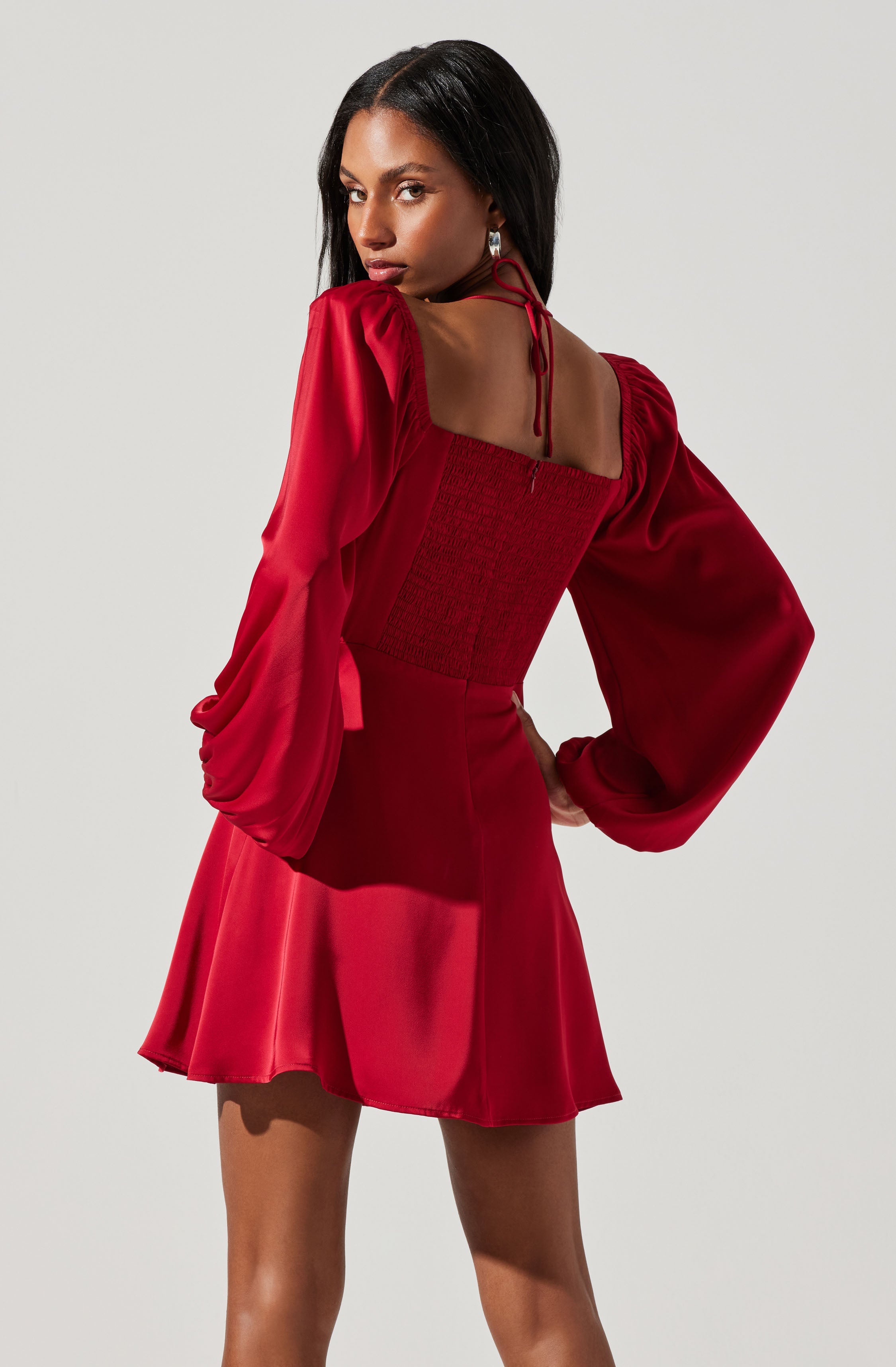 A woman wearing a red dress with long, puffed sleeves that are gathered at the wrists. The dress has a square neckline with tie details at the shoulders and a fitted, smocked bodice. The skirt is flared and above the knee.
