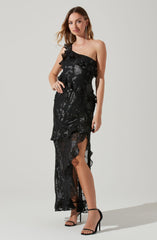 A woman is wearing a black, one-shoulder evening gown with a ruffled overlay and a slit on the side. The gown features a shiny, floral lace pattern overlaying a sheer fabric. She has on black high-heeled sandals with ankle straps and long, dangling earrings.