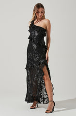 A woman is wearing a black, one-shoulder dress with ruffles and floral lace patterns. The dress features a sheer, flowy lower half with a high slit revealing her leg. She has on black high-heeled sandals with ankle straps.