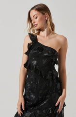 A woman is wearing a black, one-shoulder dress with ruffled, layered detailing on the strap and bodice. The fabric has a shiny, floral-patterned texture. She is accessorized with long, dangling, sparkly earrings.
