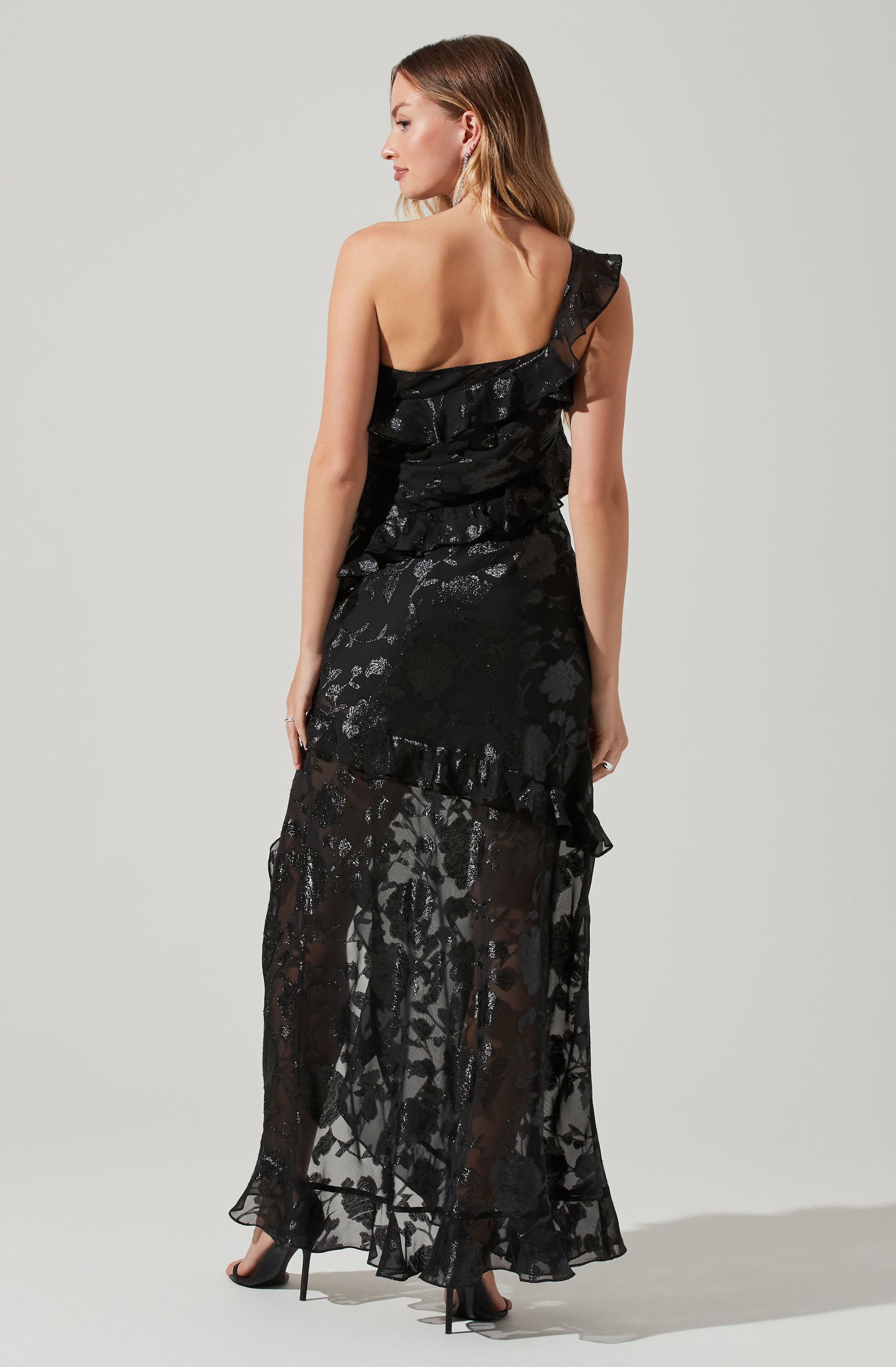 A woman is wearing a long, black dress with a sheer floral lace overlay. The dress features ruffles along the asymmetrical shoulder strap and at the waist, with a semi-transparent skirt that reveals her legs and high-heeled shoes. The lace has a shiny, metallic pattern.