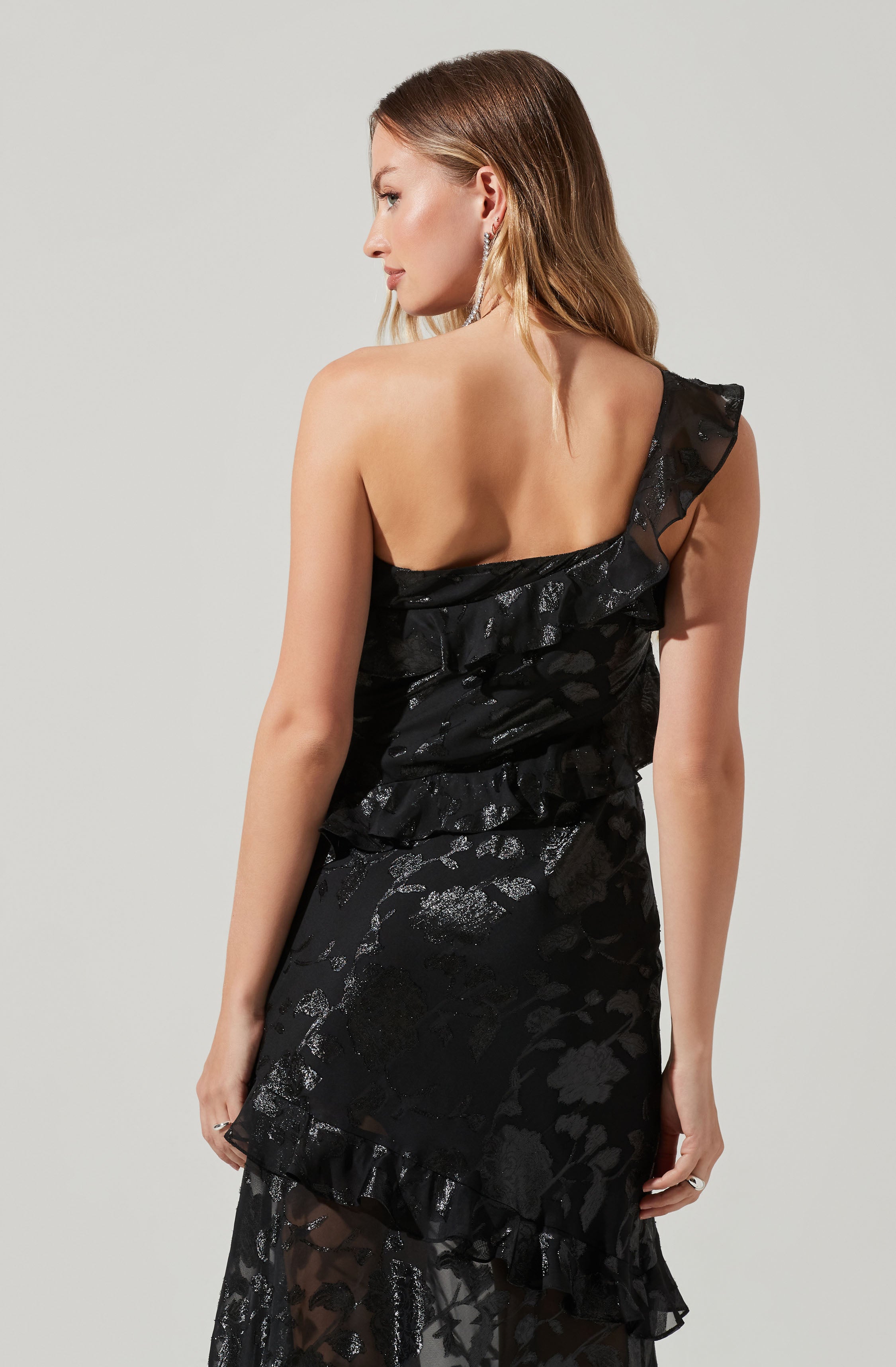 A woman is wearing a black dress with ruffled details on one shoulder. The fabric has a shiny, textured floral pattern. The dress is fitted and has a layered, ruffled hem at the bottom.