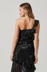 A woman is wearing a black dress with ruffled details on one shoulder. The fabric has a shiny, textured floral pattern. The dress is fitted and has a layered, ruffled hem at the bottom.