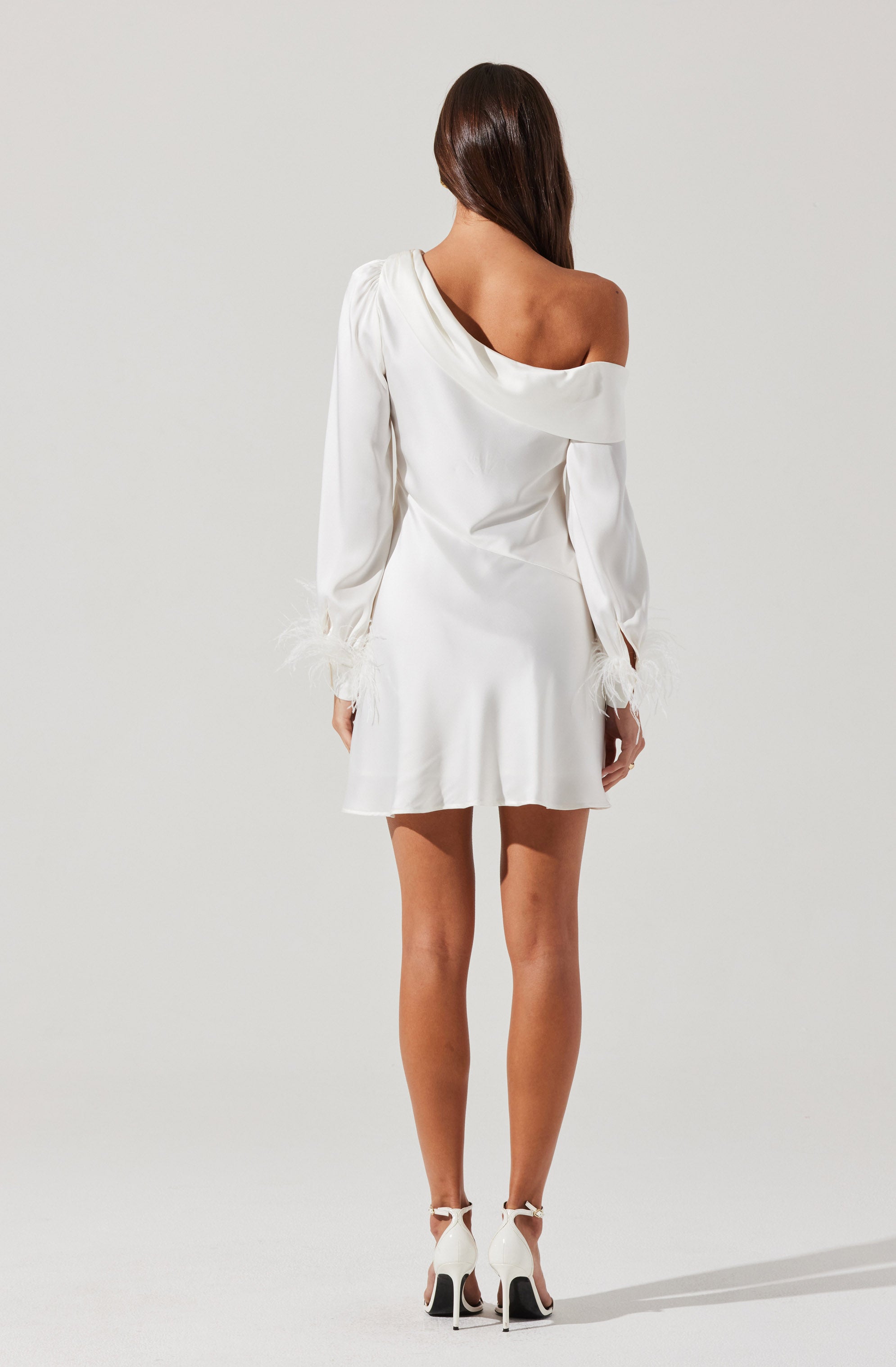 A woman is wearing a white satin dress with an off-the-shoulder neckline on the right side, exposing her shoulder and collarbone. The dress has long sleeves that gather at the wrists with feathered cuffs, and it has a slightly flared skirt that falls above the knee. She is also wearing white high-heeled shoes with ankle straps.