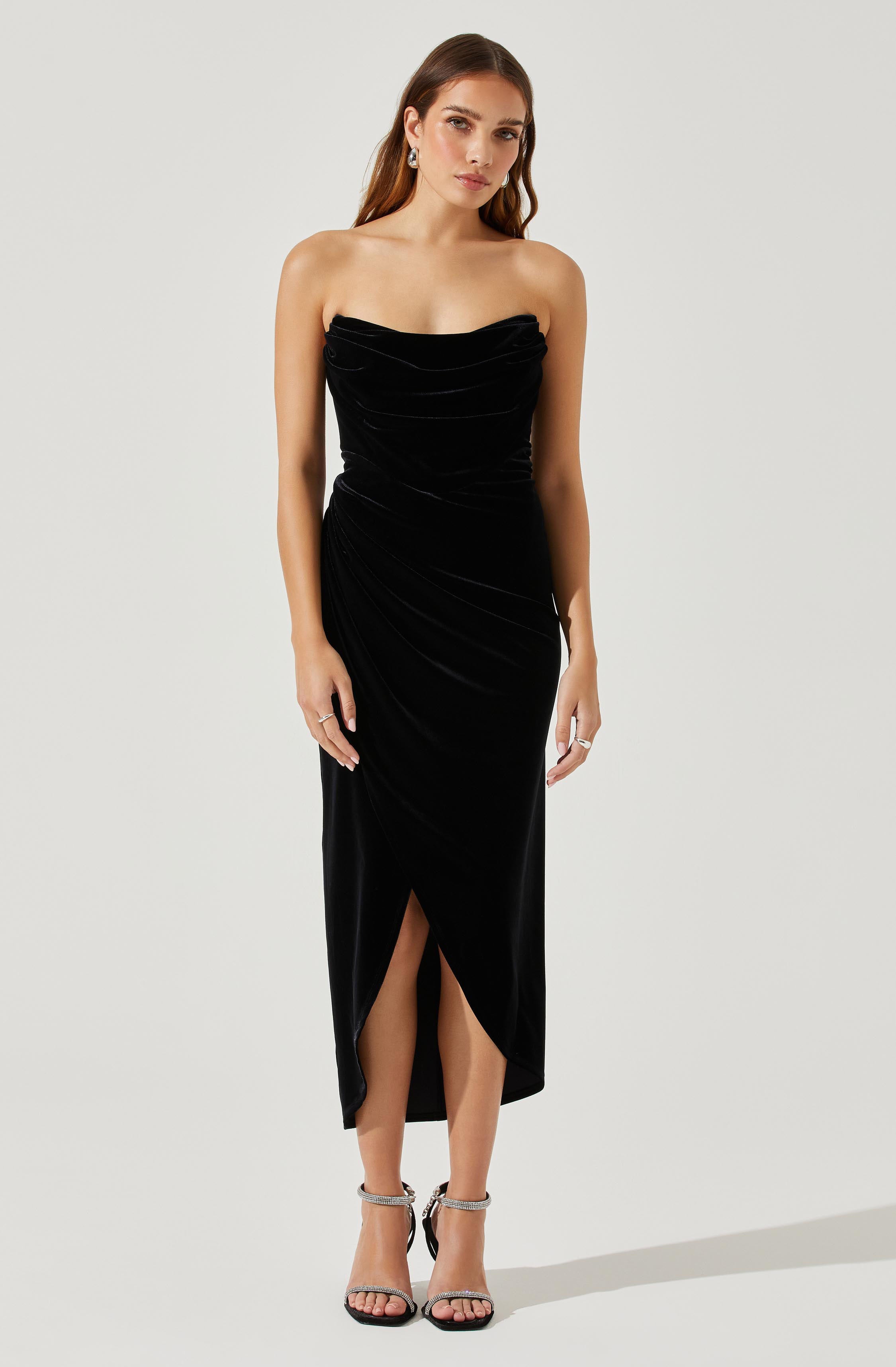 A woman is wearing a strapless black velvet dress with a ruched bodice and a high slit in the front. The dress is fitted and elegant, reaching mid-calf length. She is also wearing open-toed high-heeled sandals with ankle straps.