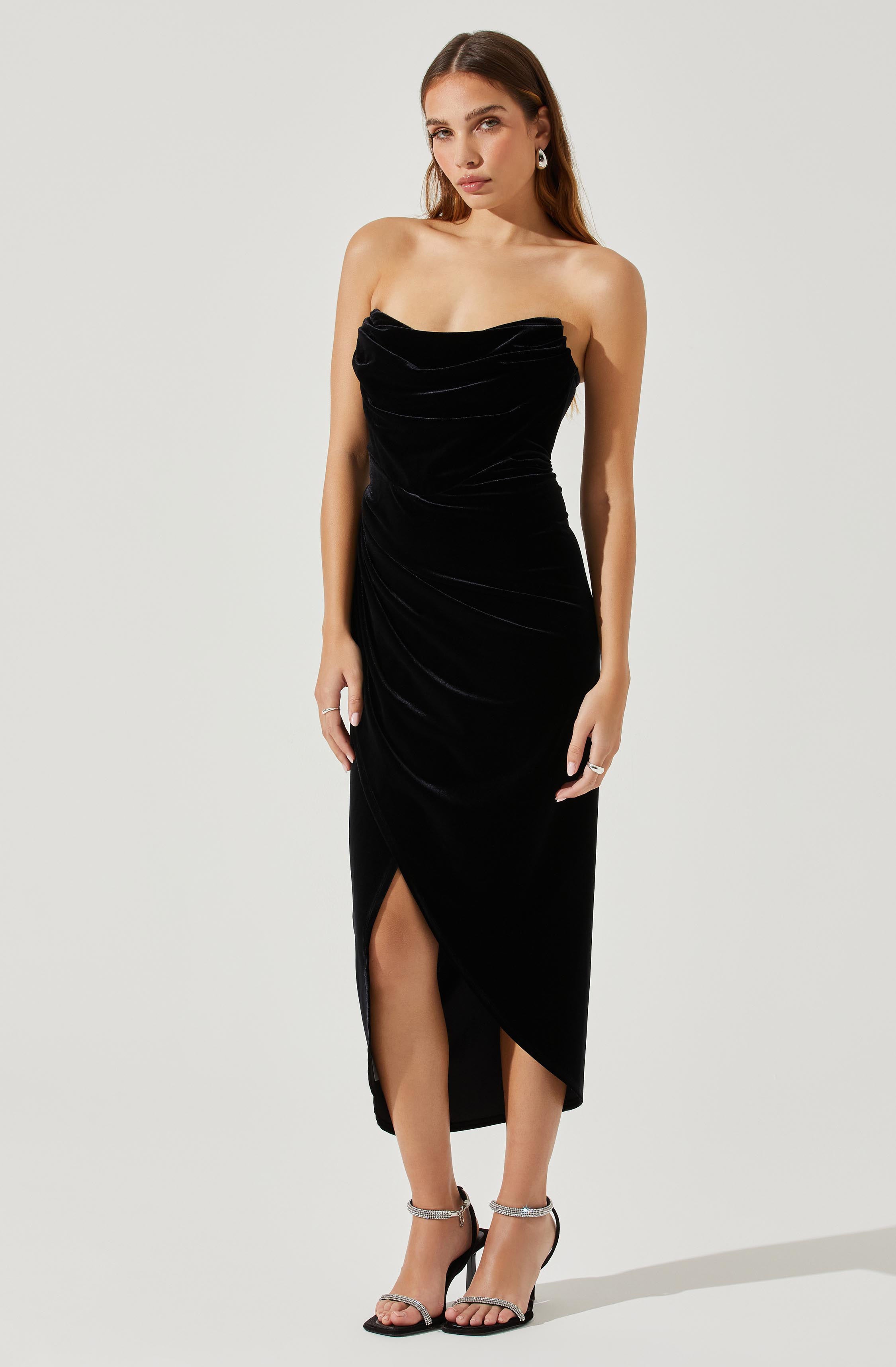 A woman is wearing a strapless, black velvet dress with a draped, ruched bodice and an asymmetrical hemline with a front slit. She has on silver ankle-strap high-heeled sandals and silver hoop earrings.