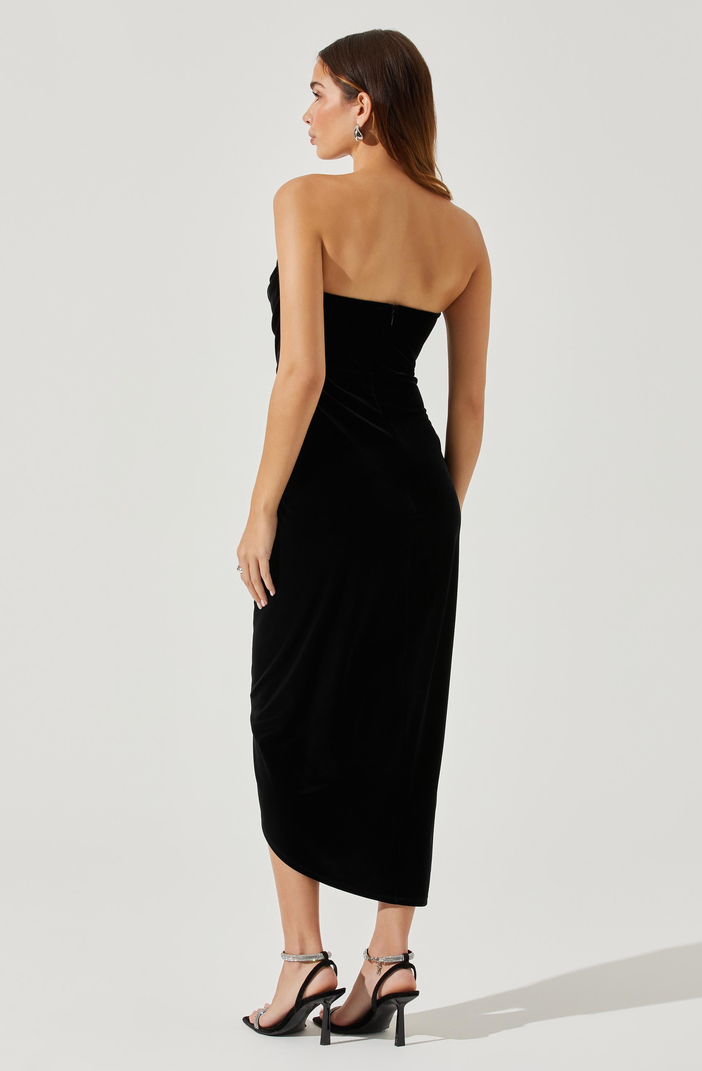 A woman is wearing a strapless, floor-length black dress made of a shiny, velvety material. The dress is fitted through the bodice and hips, and has a slightly gathered detail on one side. She is also wearing black high-heeled sandals with ankle straps.
