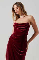 A woman is wearing a strapless dress made of deep red velvet fabric. The dress features a ruched bodice with gathered detailing around the bust and waist, creating a draped effect. The fabric extends down in elegant folds, and there is a subtle slit on one side of the dress.