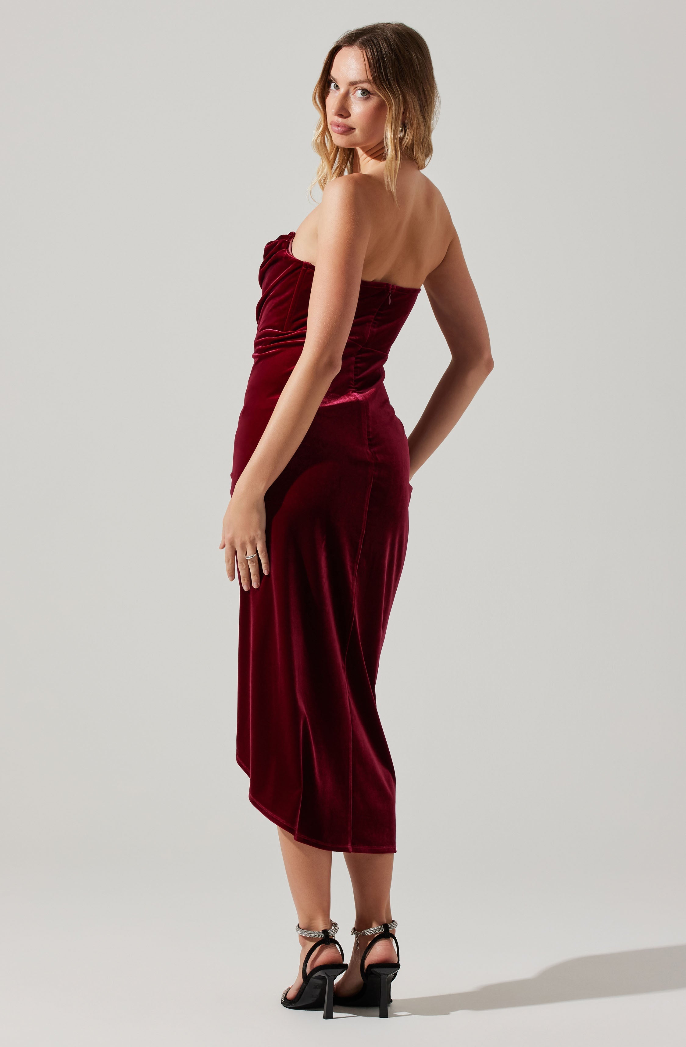 A woman wearing a strapless, deep red velvet dress that is fitted through the bodice and hips, with a draped detail at the top. The dress has a straight, mid-length hemline that falls below the knees. She is also wearing high-heeled sandals with ankle straps.