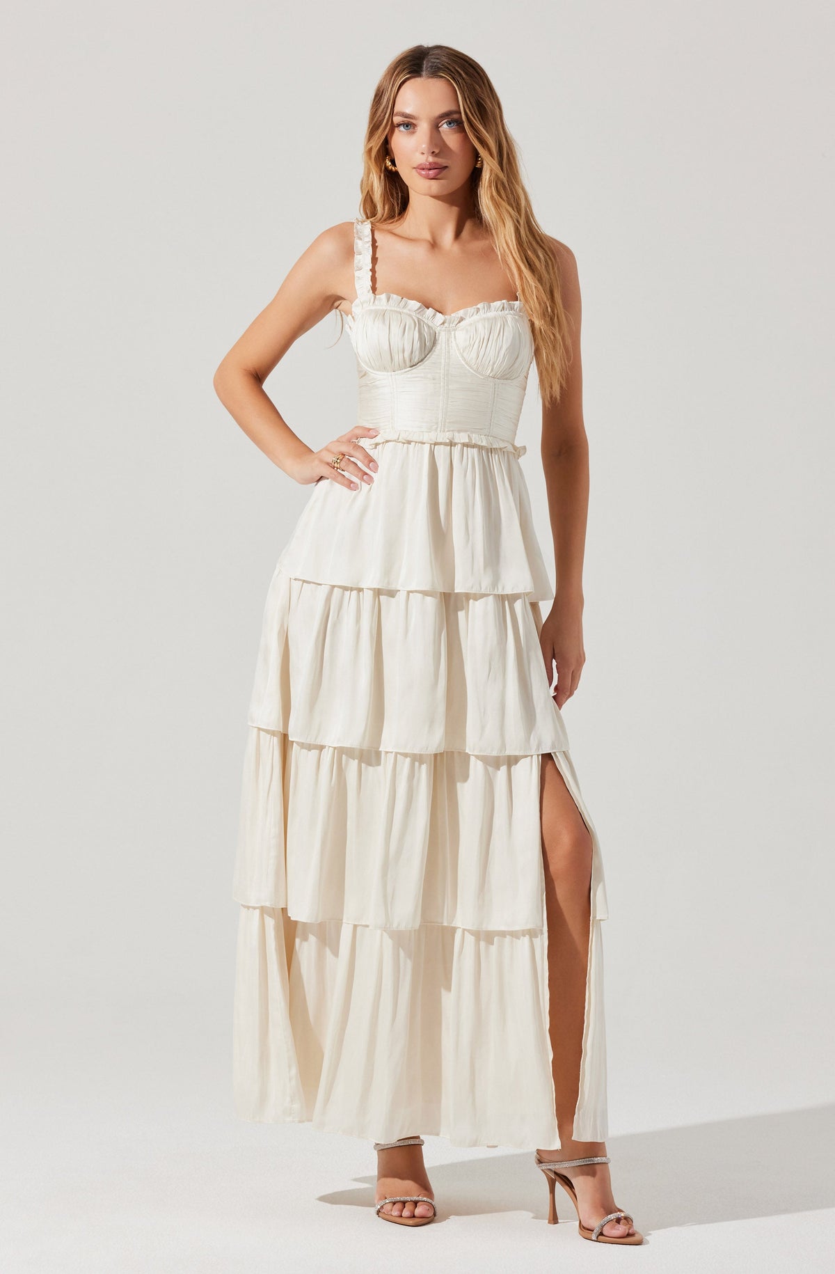 A woman is wearing a cream-colored, sleeveless maxi dress with ruffled shoulder straps. The dress has a fitted bodice with subtle textured detailing and a tiered, flowing skirt with a high slit on one side. She is also wearing nude high-heeled sandals.