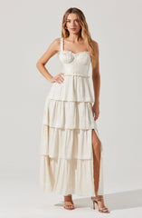 A woman is wearing a cream-colored, sleeveless maxi dress with ruffled shoulder straps. The dress has a fitted bodice with subtle textured detailing and a tiered, flowing skirt with a high slit on one side. She is also wearing nude high-heeled sandals.