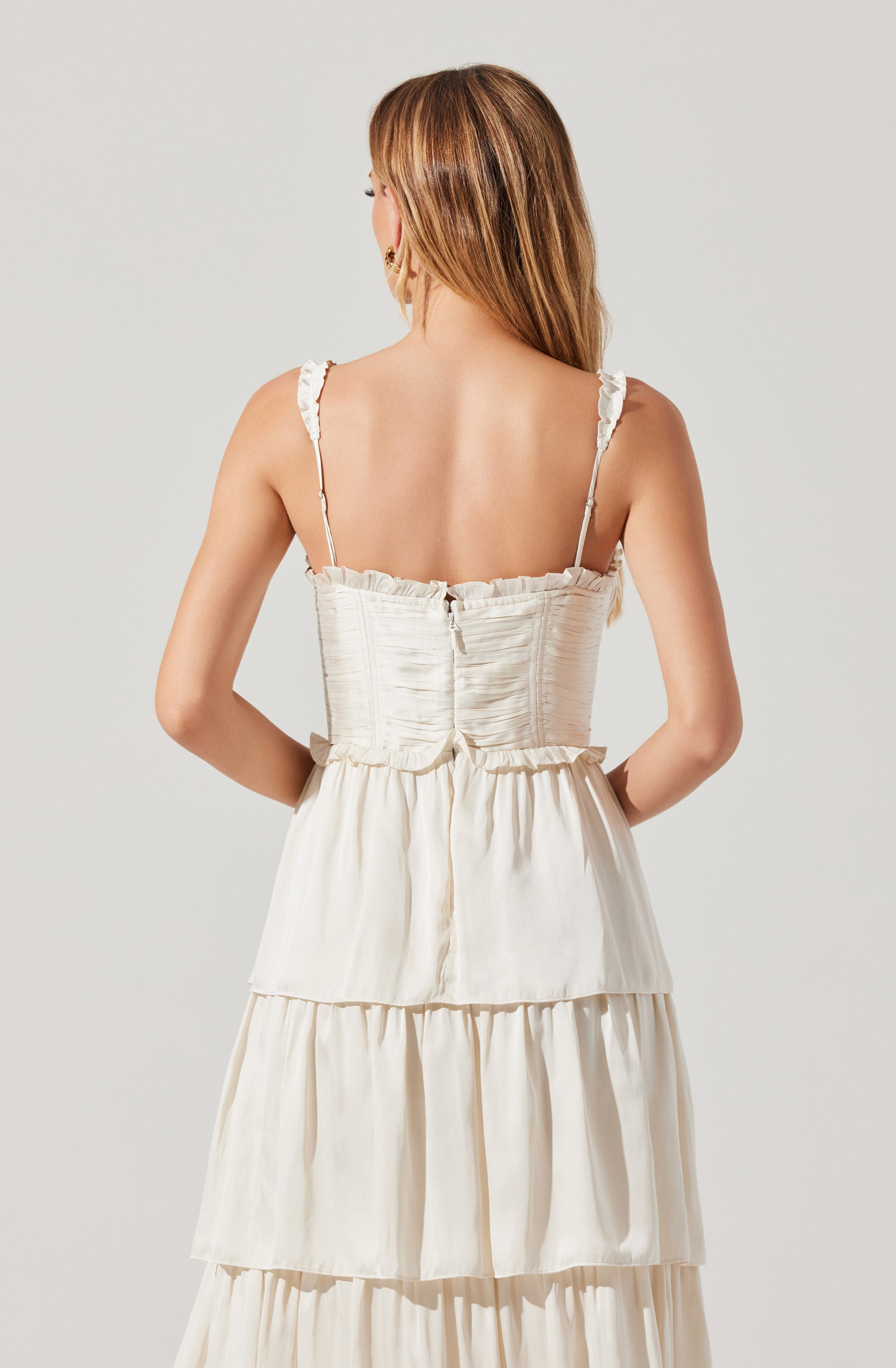A woman is wearing a sleeveless, off-white, tiered dress with ruffle shoulder straps. The dress has a fitted, ruched bodice with a back zipper. The skirt features multiple gathered layers, creating a flowing, multi-tiered look.
