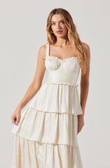 A woman is wearing a sleeveless, ivory-colored dress with a fitted bodice featuring ruched detailing and slightly ruffled straps. The dress has a tiered, flowy skirt with multiple layers creating a ruffled appearance.