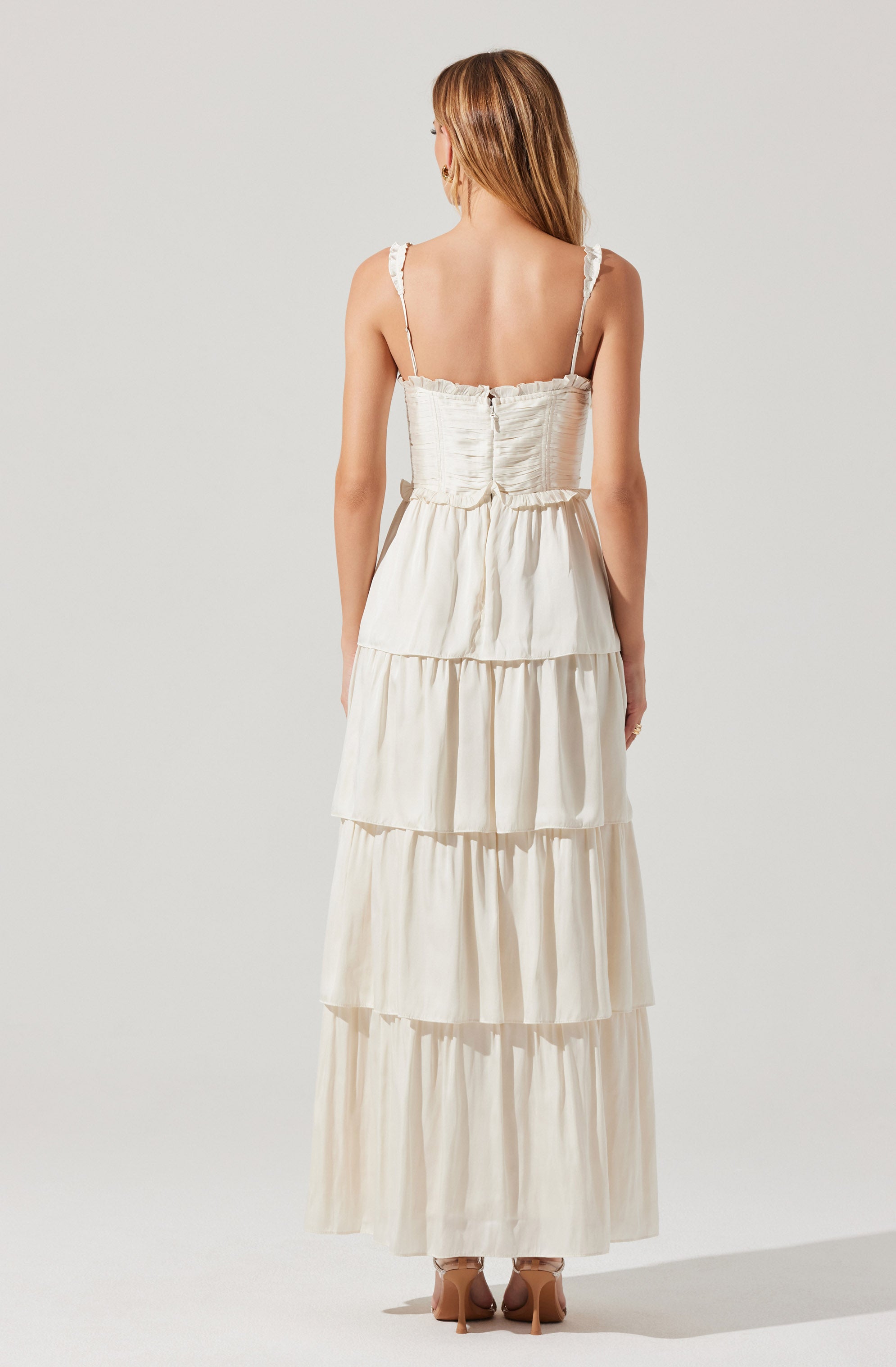 A woman is wearing a sleeveless, tiered, cream-colored maxi dress with ruffled straps and a fitted, ruched bodice that has a zipper in the back. The dress features multiple horizontal layers with gathered fabric, creating a flowing silhouette. She is standing against a plain, light-colored background.