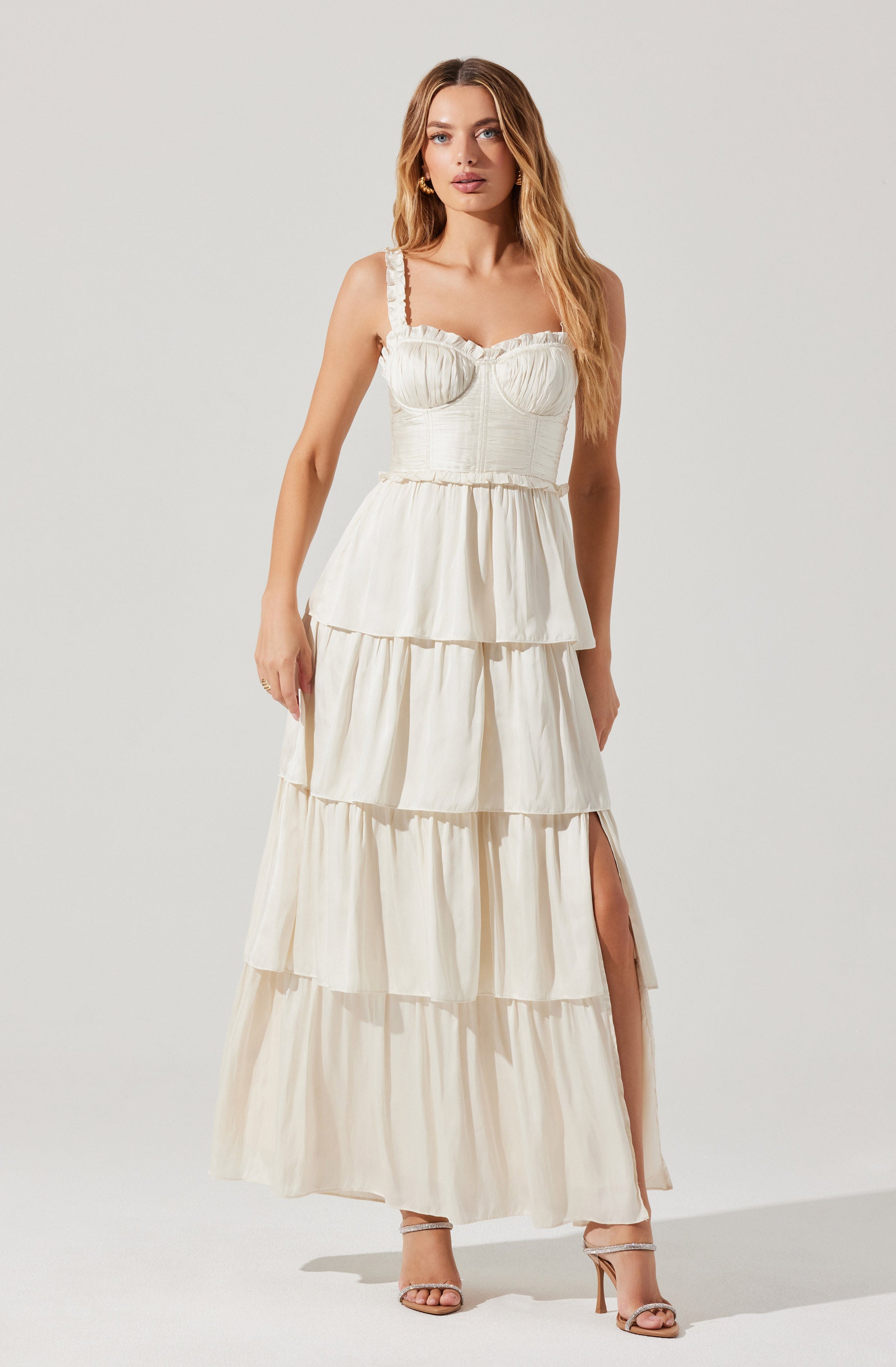 A woman is wearing a sleeveless, white tiered maxi dress with ruffled straps and a fitted bodice featuring a sweetheart neckline. The dress has multiple horizontal tiers with gathered fabric, creating a flowing, voluminous skirt with a side slit.