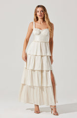 A woman is wearing a sleeveless, white tiered maxi dress with ruffled straps and a fitted bodice featuring a sweetheart neckline. The dress has multiple horizontal tiers with gathered fabric, creating a flowing, voluminous skirt with a side slit.