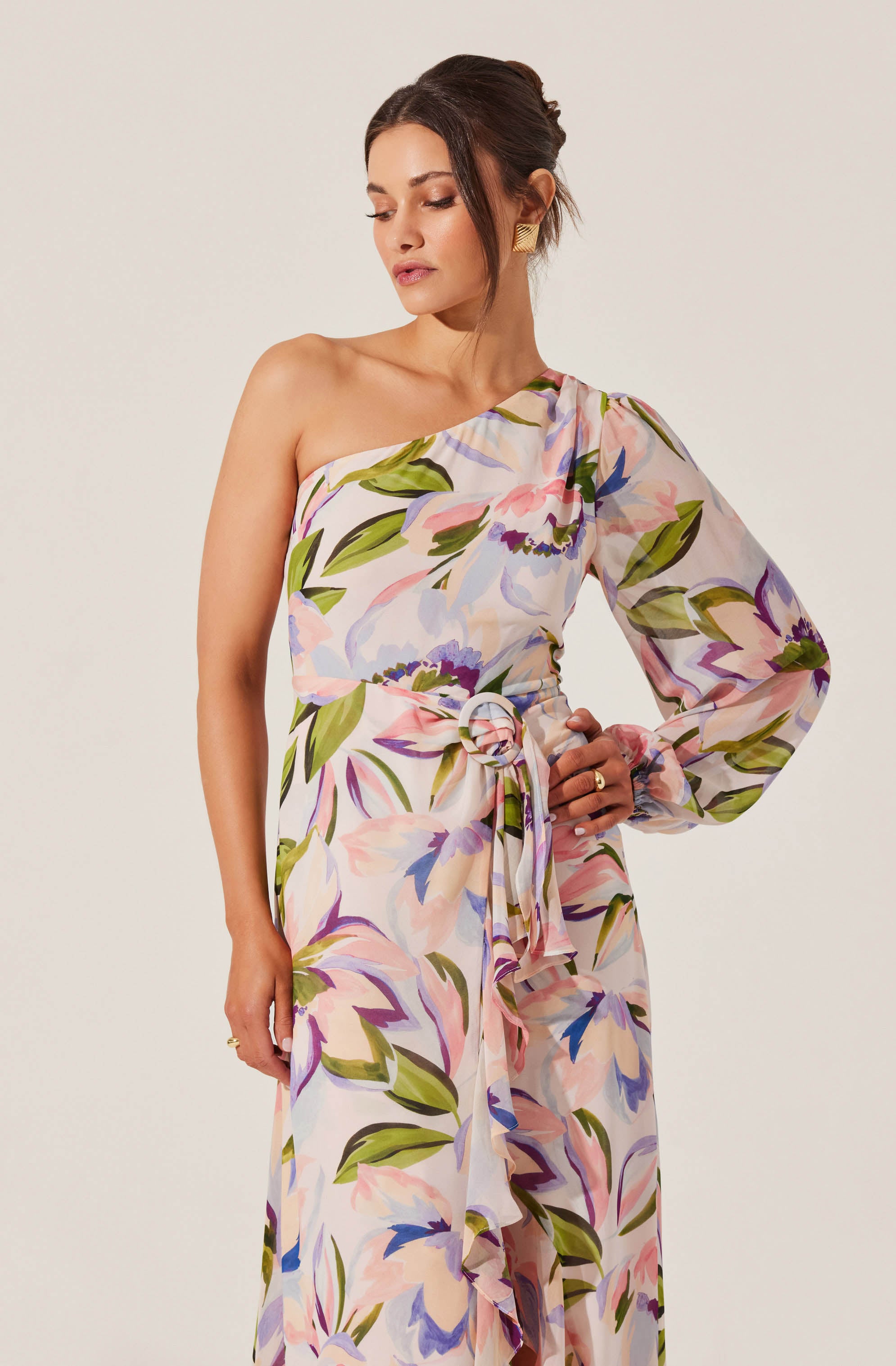 A woman is wearing a one-shoulder dress with a floral pattern featuring large, colorful flowers and green leaves. The dress has a long, flowing skirt and a fabric belt tied in a knot at the waist. The fabric appears lightweight and semi-sheer, with a soft, pastel color palette including shades of pink, purple, blue, and green.