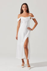 A woman is wearing a white, off-the-shoulder dress with spaghetti straps. The dress features ruffled details on the shoulders and a thigh-high slit on one side. She is also wearing white heels with pearl embellishments.