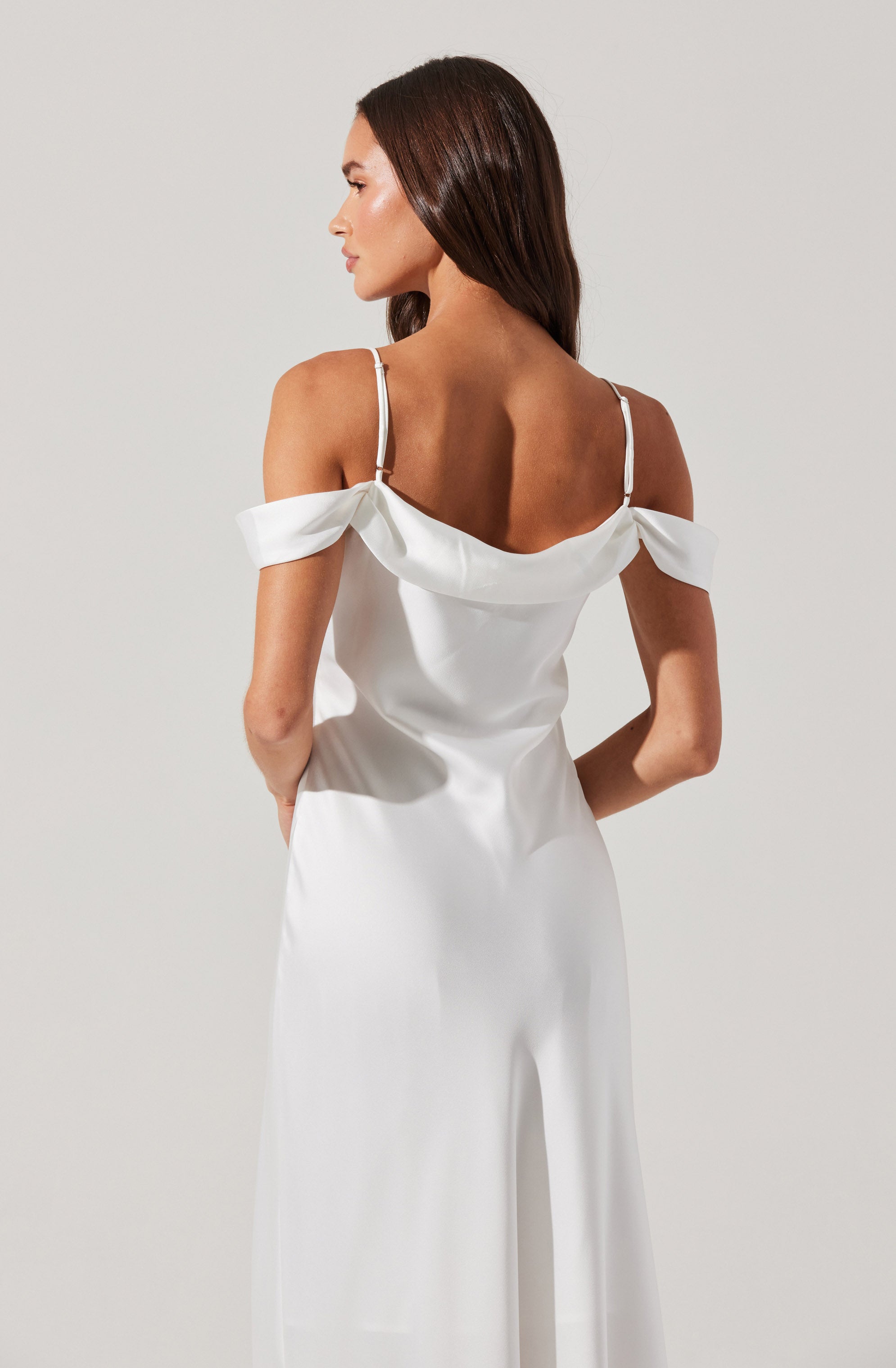 A woman is wearing a white satin jumpsuit with off-the-shoulder sleeves that are draped and gathered. The jumpsuit features thin shoulder straps and has a smooth, flowing appearance from the fitted waist down to wide-leg trousers.