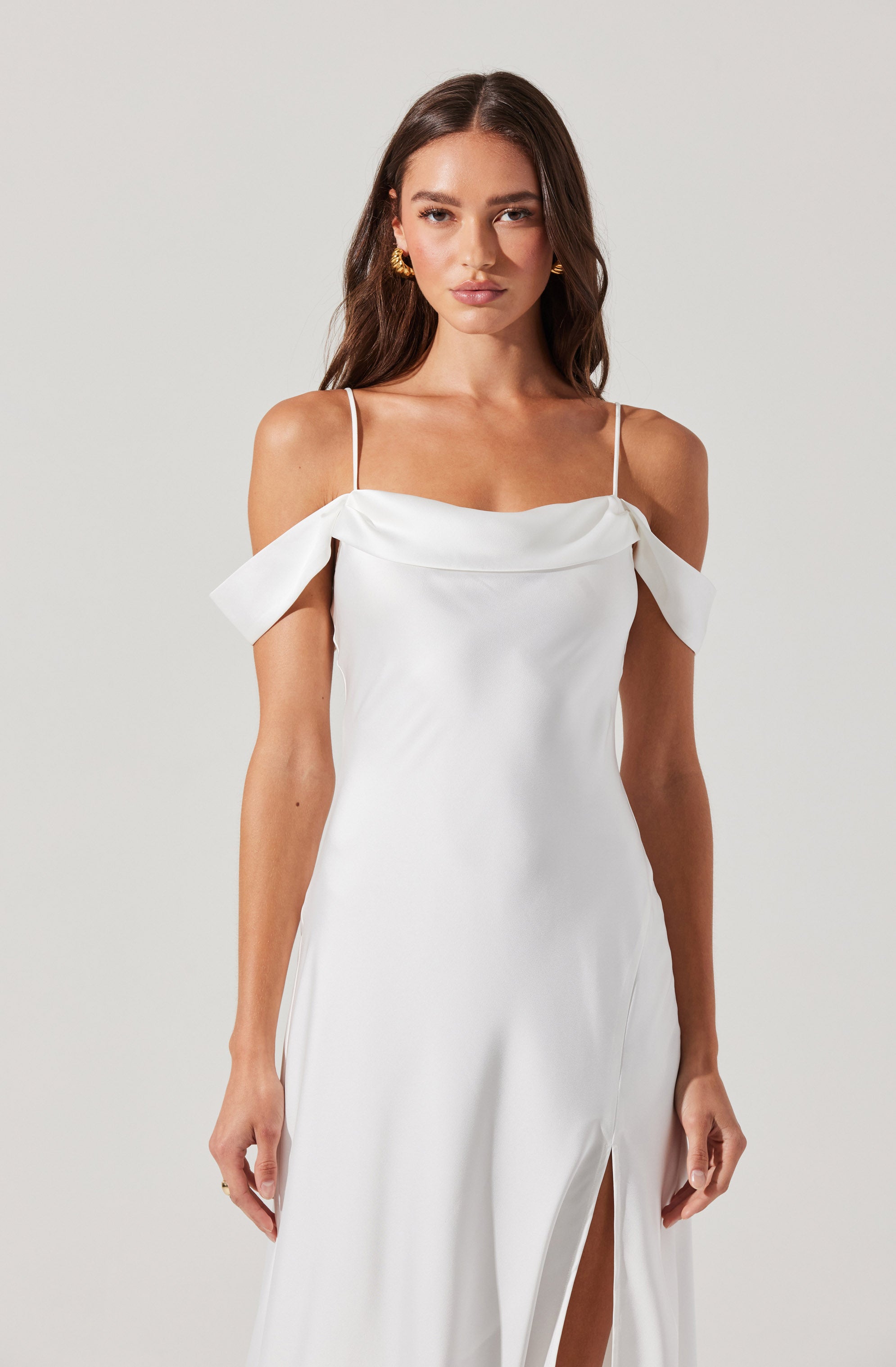 A woman is wearing a white gown with thin spaghetti straps. The dress features off-the-shoulder ruffled sleeves and a high slit in the front, revealing her legs.