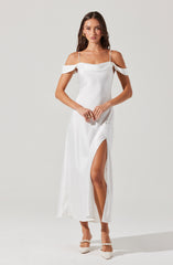 A woman is wearing a white, off-the-shoulder dress with thin spaghetti straps. The dress features ruffled shoulder details and a high slit on the left side, revealing her leg. She is also wearing white shoes with ankle straps decorated with pearls.
