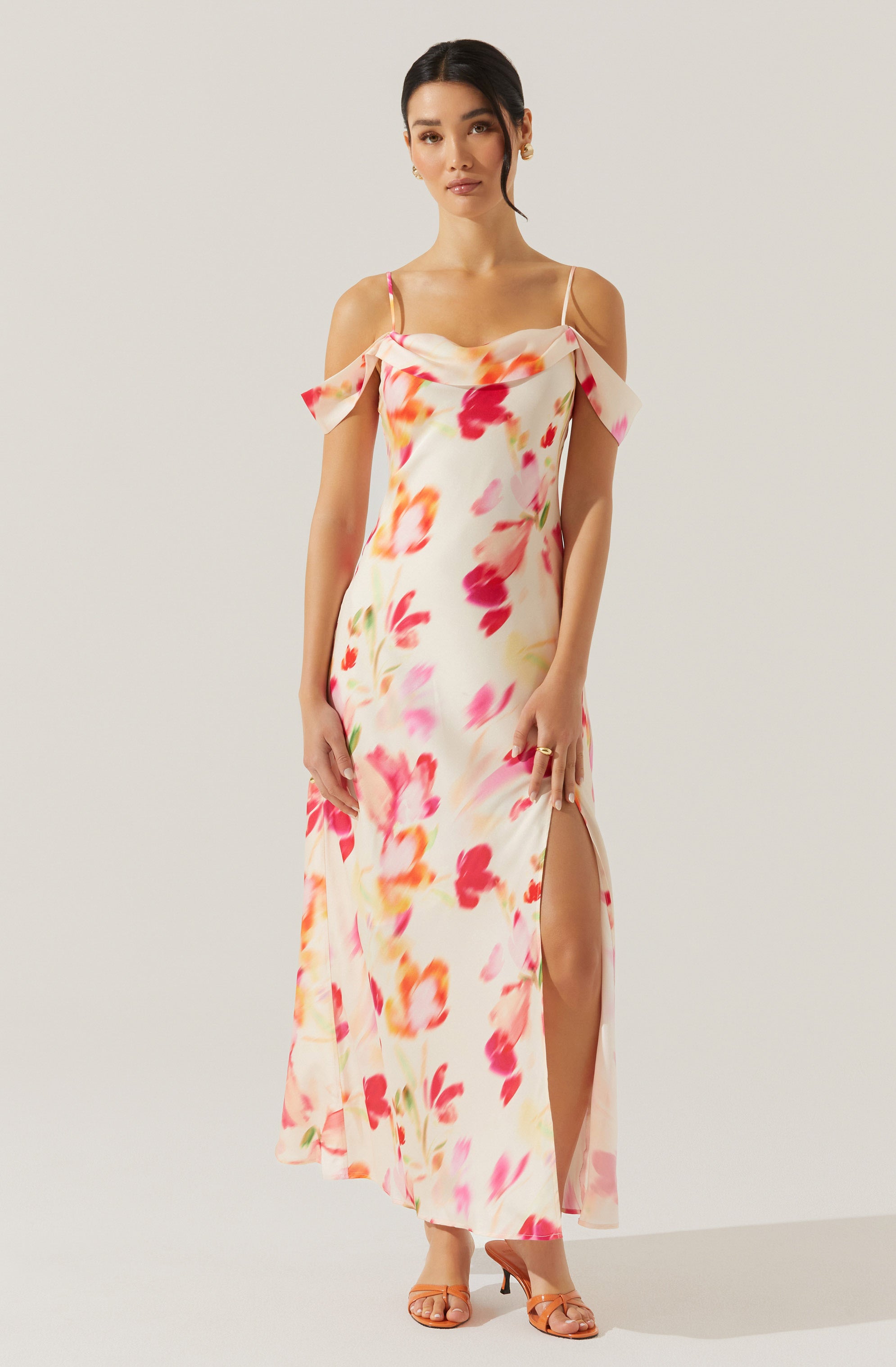 A woman wearing a long, flowy dress with a floral pattern featuring shades of pink, red, orange, and yellow. The dress has thin spaghetti straps, off-the-shoulder flutter sleeves, and a high slit on one side.
