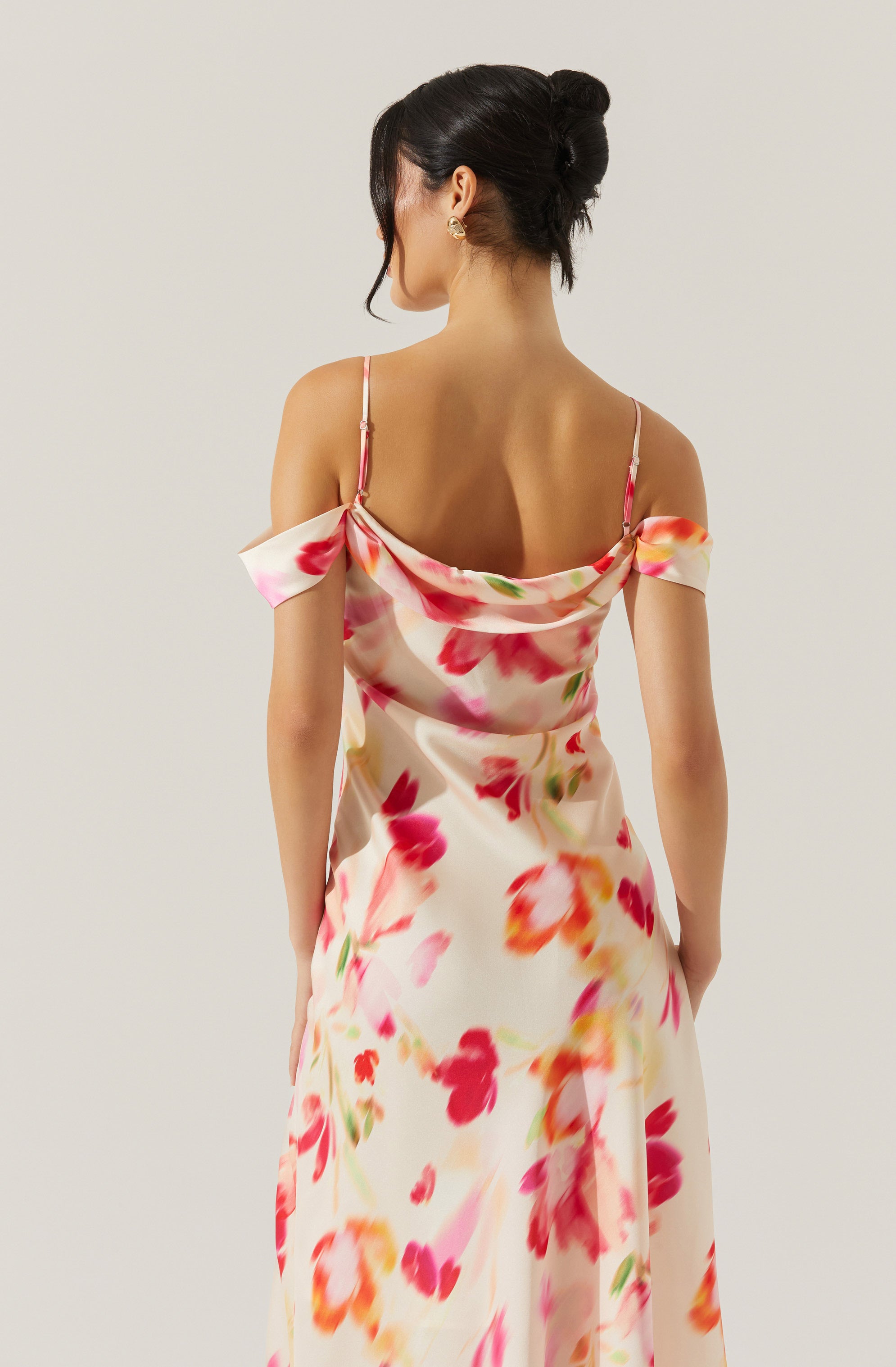 A woman is wearing a silky, floral dress with a cream background and vibrant pink, red, orange, and green flower patterns. The dress features spaghetti straps and off-the-shoulder sleeves. The fabric has a smooth, shiny texture.
