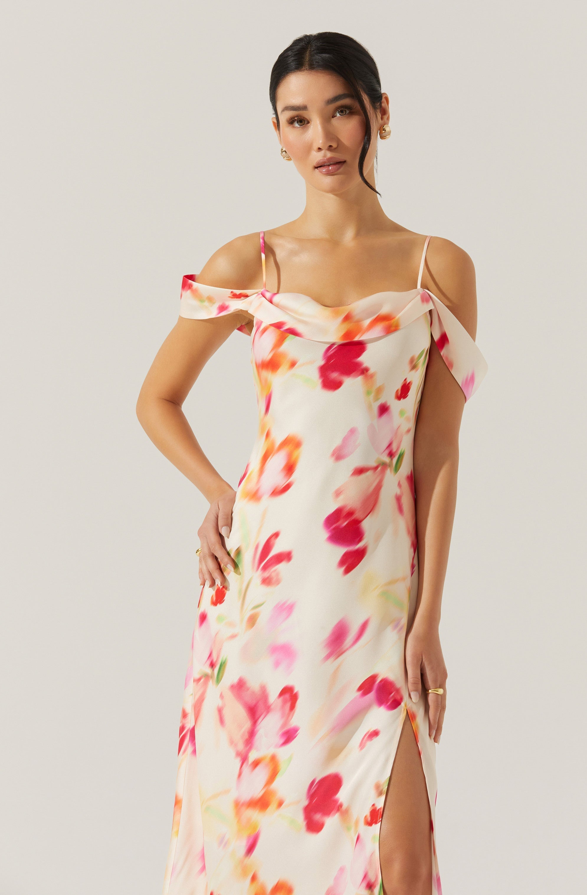 A woman is wearing a sleeveless, floor-length dress with a floral pattern in shades of pink, red, orange, and green on a light cream background. The dress features a draped off-the-shoulder neckline with thin spaghetti straps and a high slit on the side.