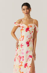A woman is wearing a sleeveless, floor-length dress with a floral pattern in shades of pink, red, orange, and green on a light cream background. The dress features a draped off-the-shoulder neckline with thin spaghetti straps and a high slit on the side.