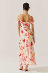 A woman is wearing a long, flowy dress with a colorful floral pattern featuring shades of pink, orange, and red on a light, creamy background. The dress has off-the-shoulder sleeves with small bows and thin spaghetti straps.