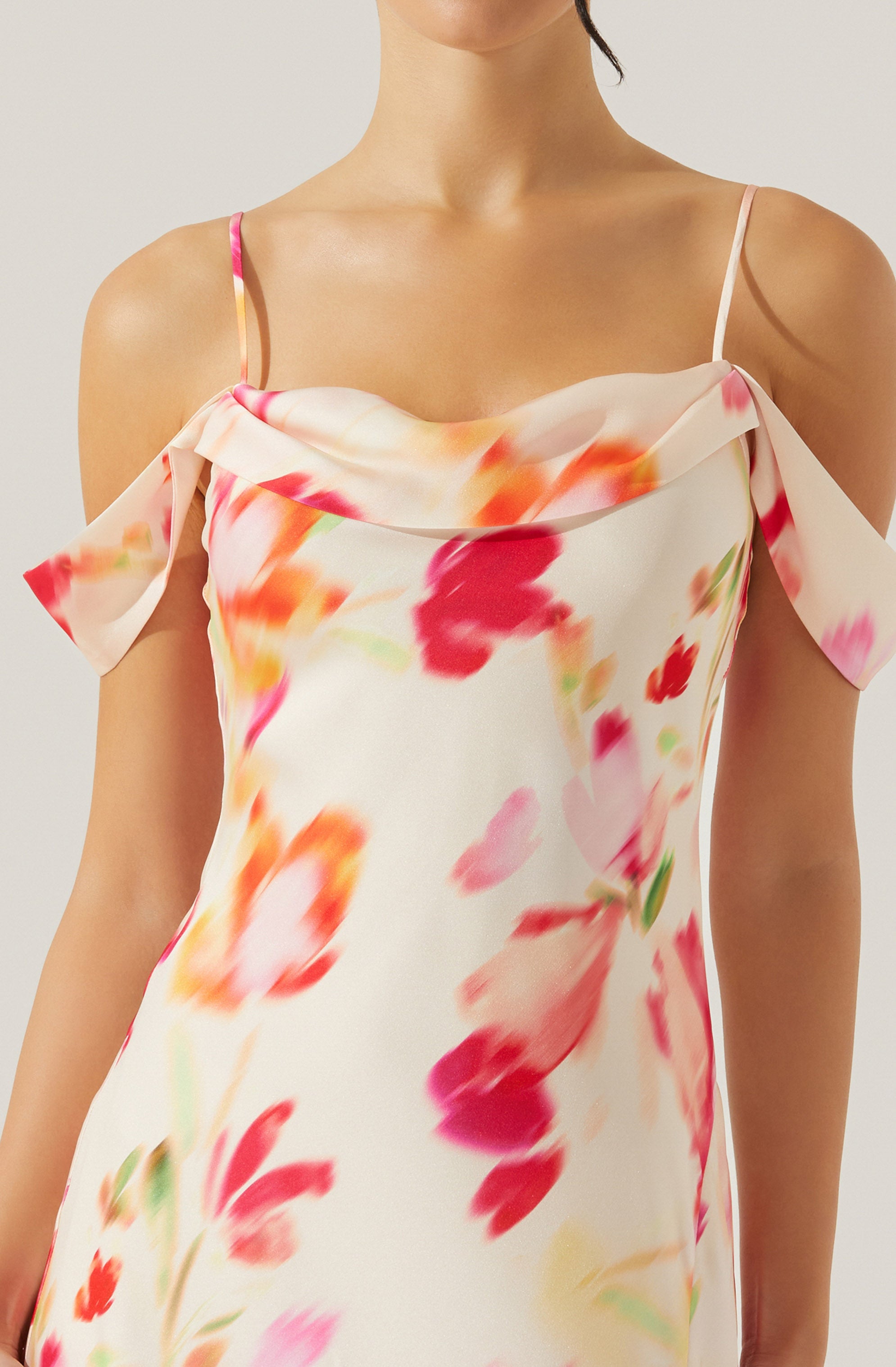 A woman is wearing a sleeveless, off-the-shoulder dress with thin spaghetti straps. The dress has a soft, flowing fabric with a floral pattern featuring large, blurred flowers in shades of pink, red, orange, and green on a light, creamy background. The neckline drapes loosely across the shoulders, creating a relaxed and elegant look.